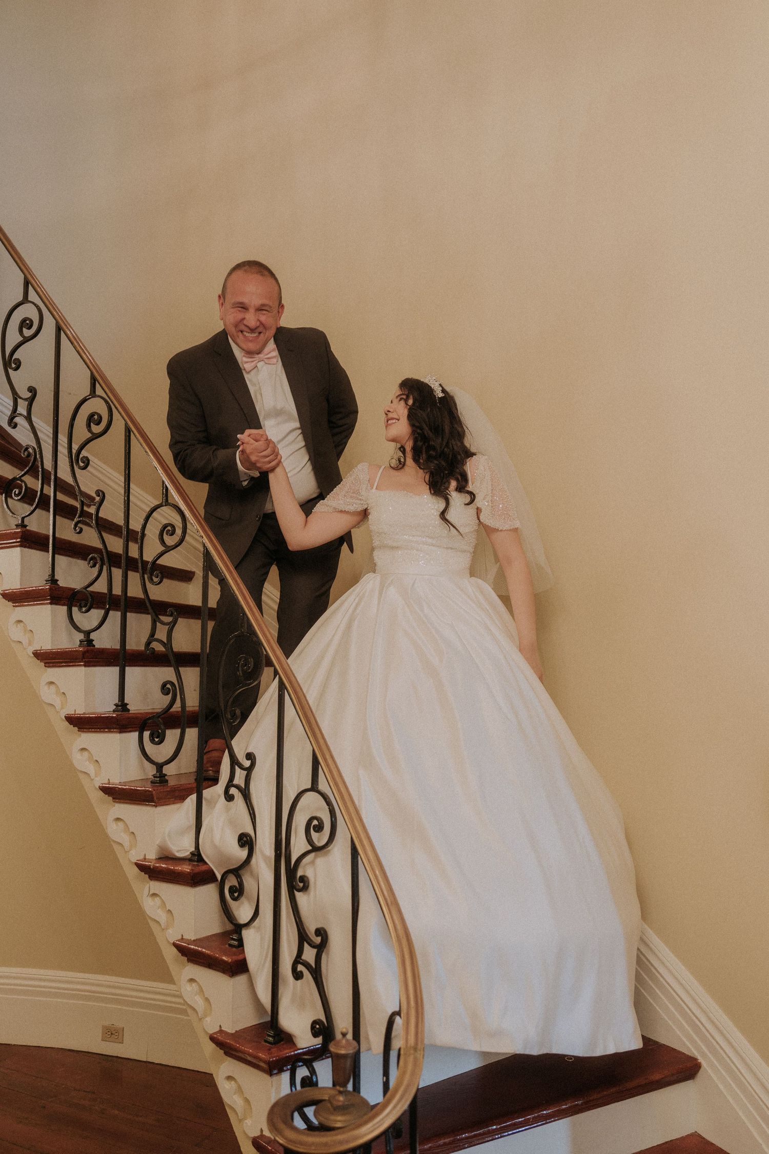 Bride and groom holding hands on elegant spiral staircase, bride in white ball gown, groom in dark suit, smiling.