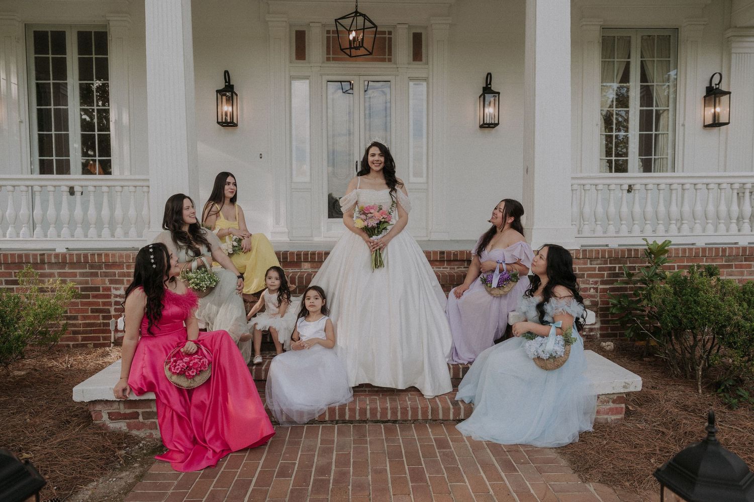 Bride in white gown poses with colorful bridesmaids and flower girls on brick steps of elegant white mansion.