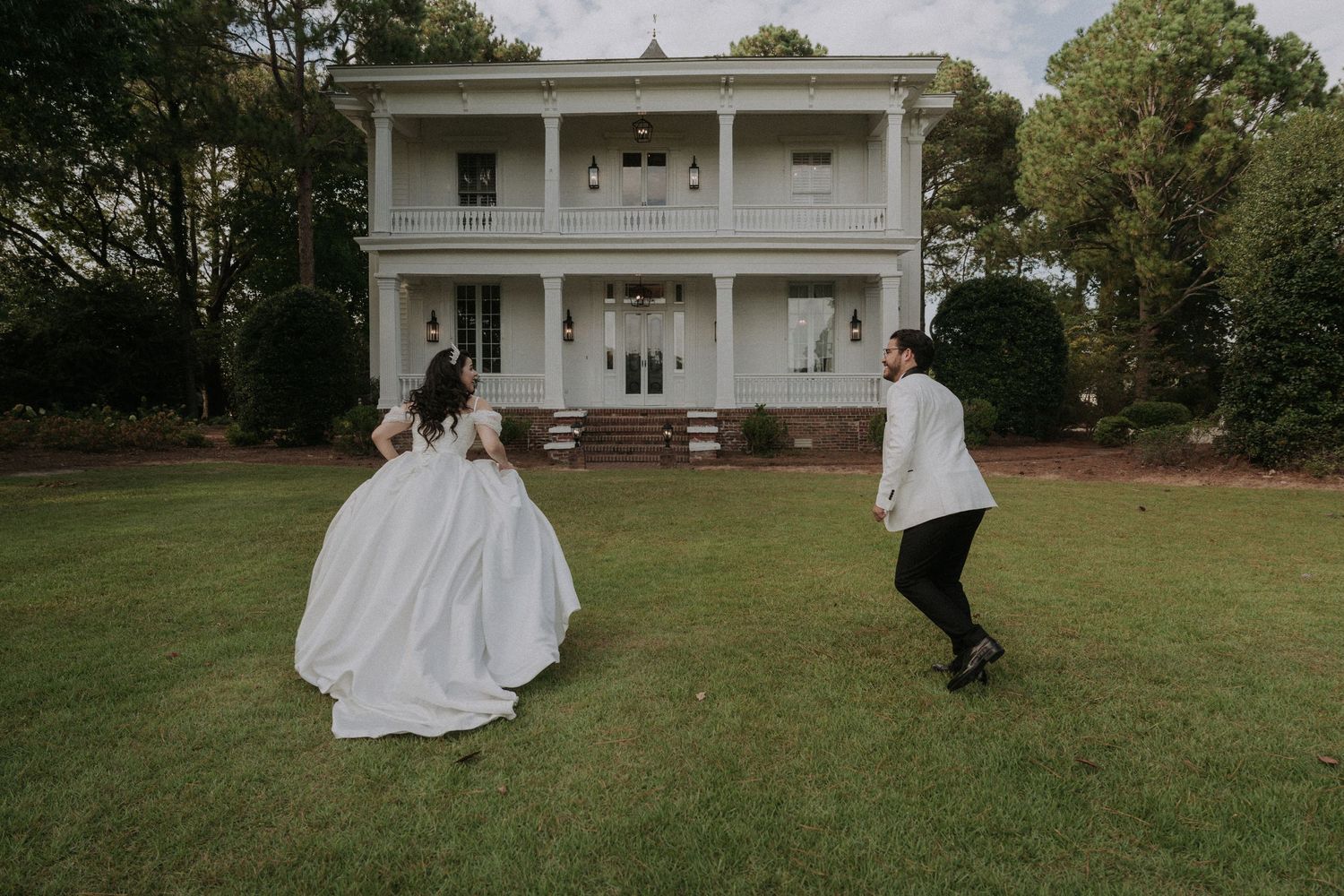 Bride and groom running across green lawn toward elegant white two-story plantation-style wedding venue.