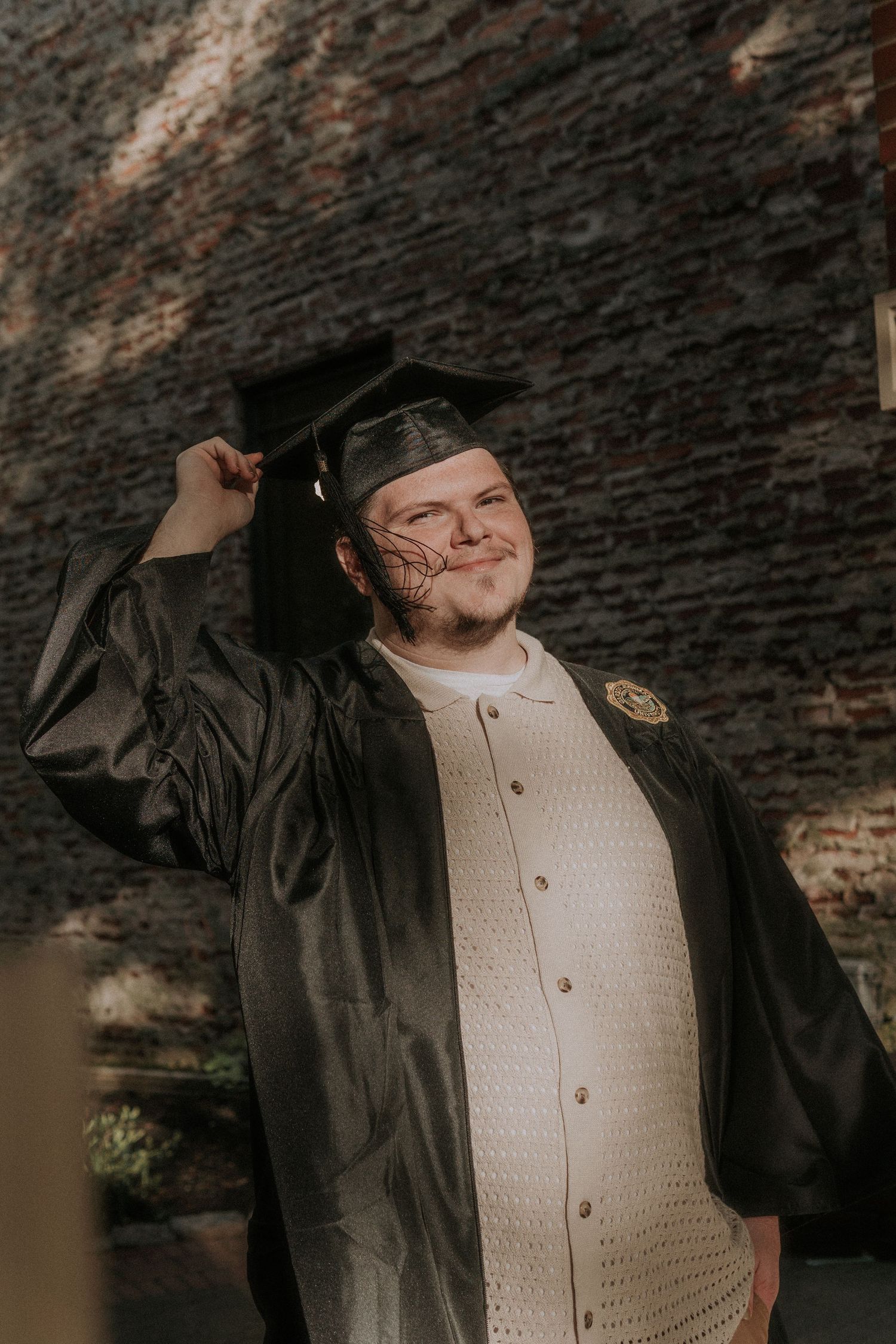 A male graduate in cap and gown poses against a brick wall with a joyful expression.