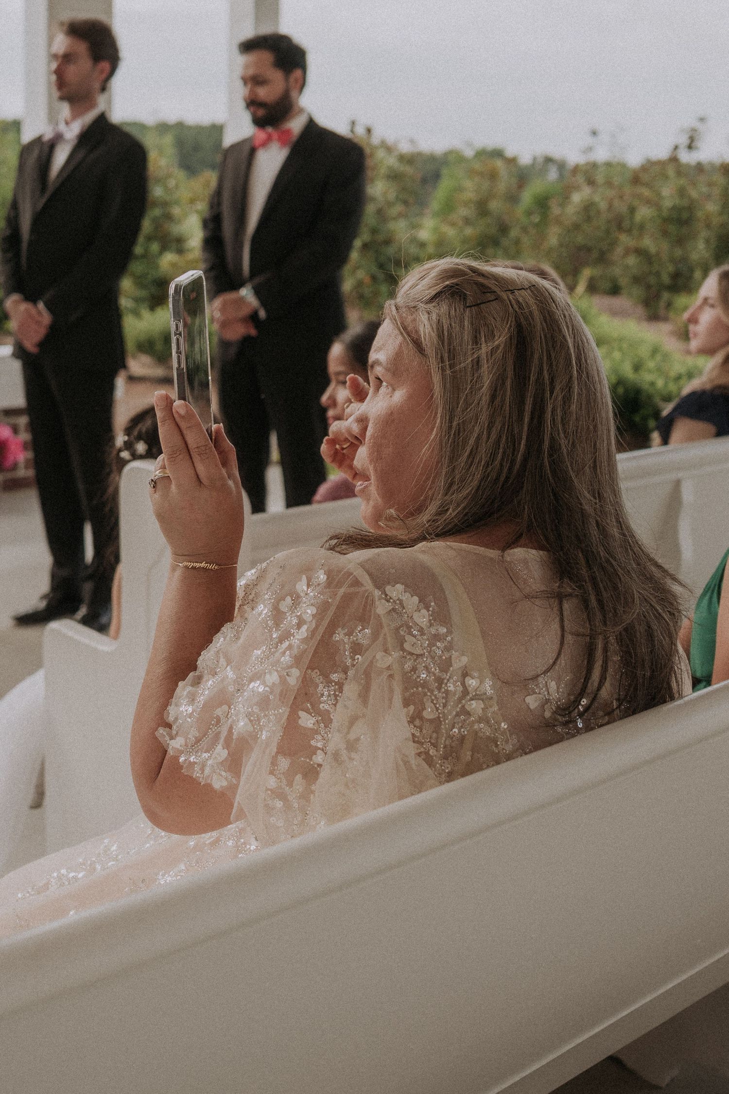 A wedding guest in lace cape dress photographs the ceremony on her phone while seated in white pews.