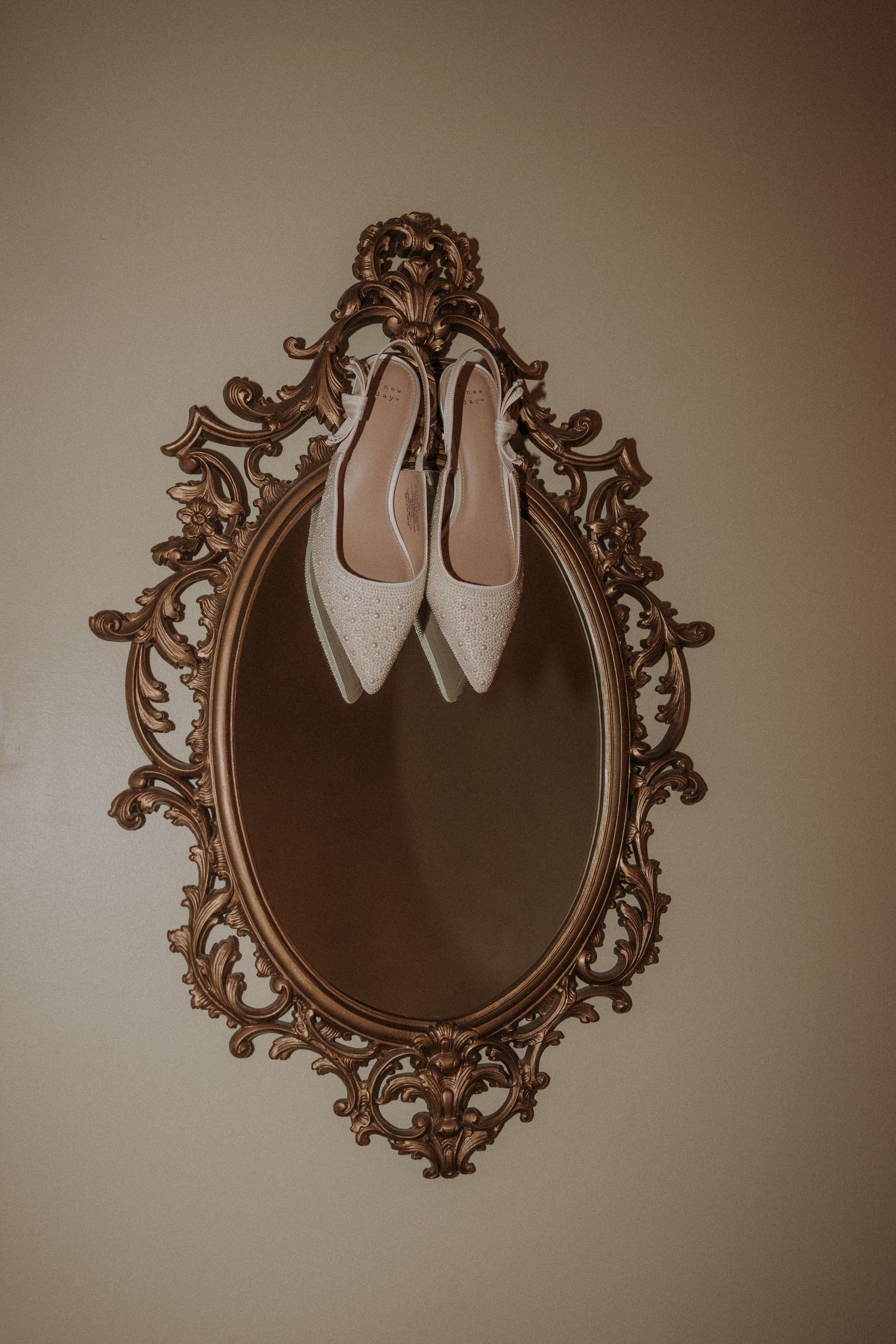 White glittery bridal heels displayed on an ornate vintage gold oval mirror hanging on a beige wall.