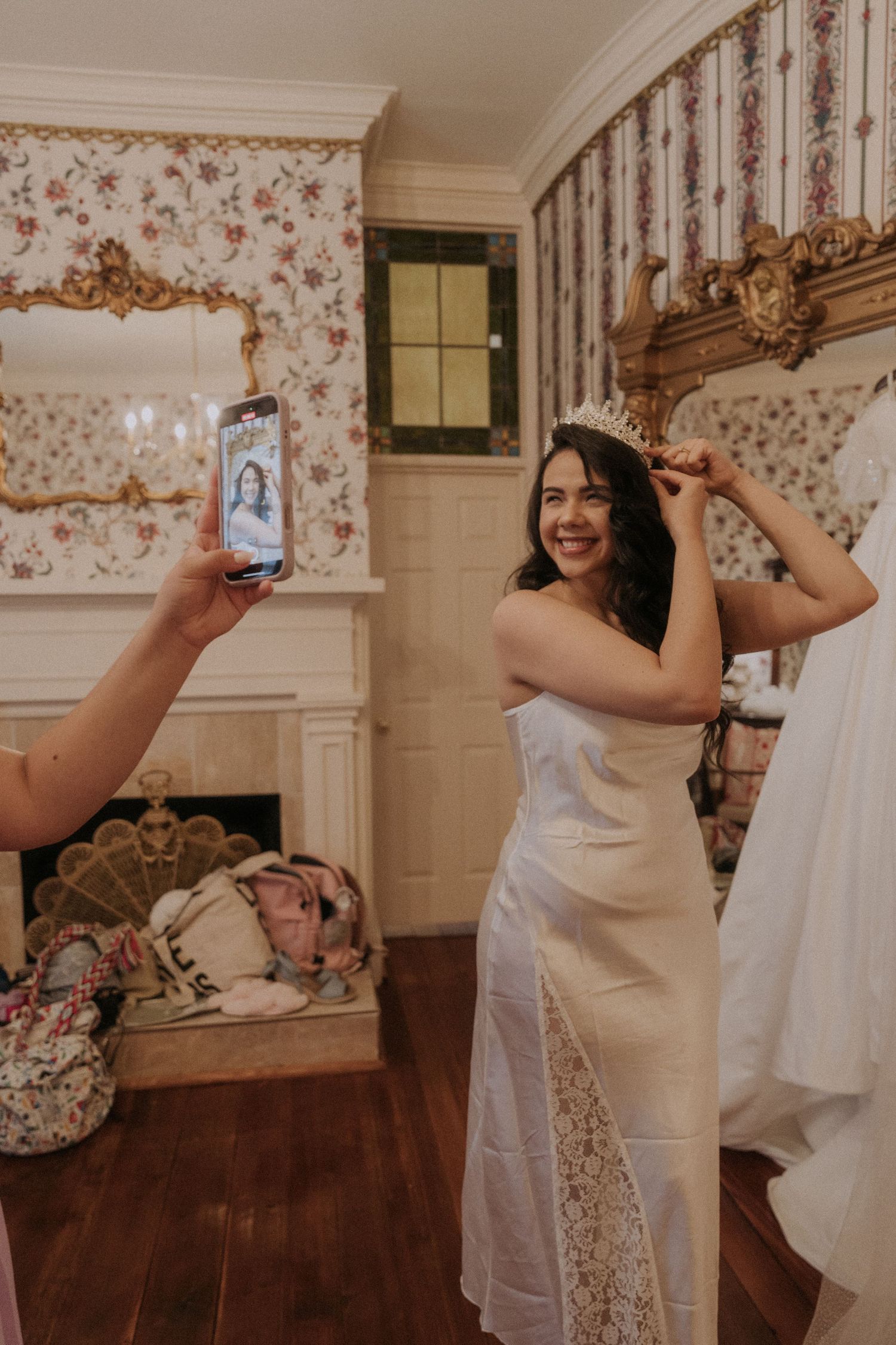 Smiling bride in white wedding dress being photographed while placing tiara on head in vintage room.