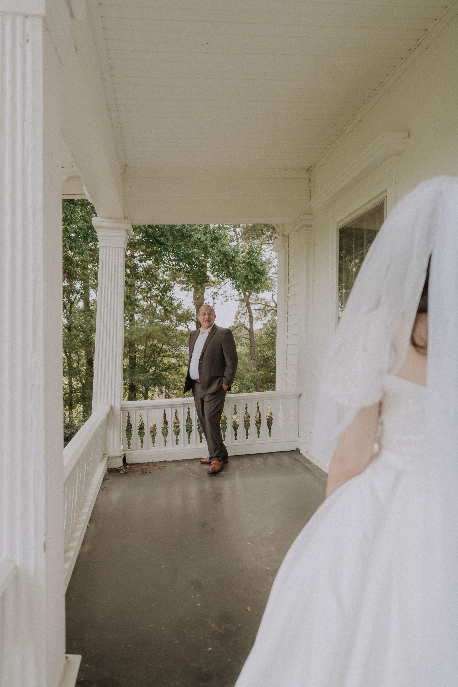 Groom smiling at bride in white wedding dress and veil on elegant white porch with columns.