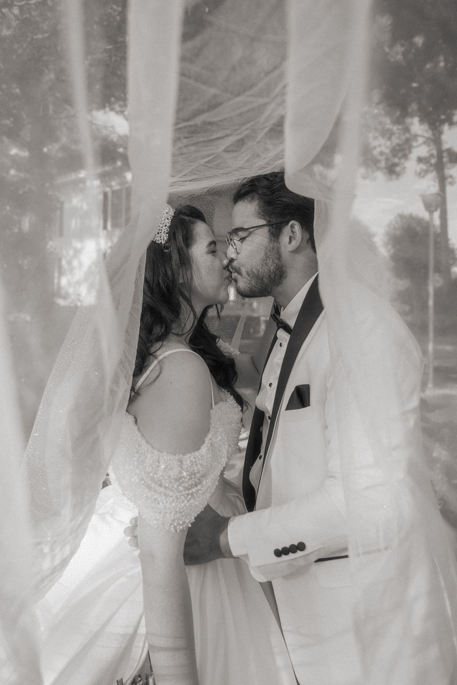 Black and white photo of a bride and groom sharing a kiss under flowing white fabric at their wedding ceremony.