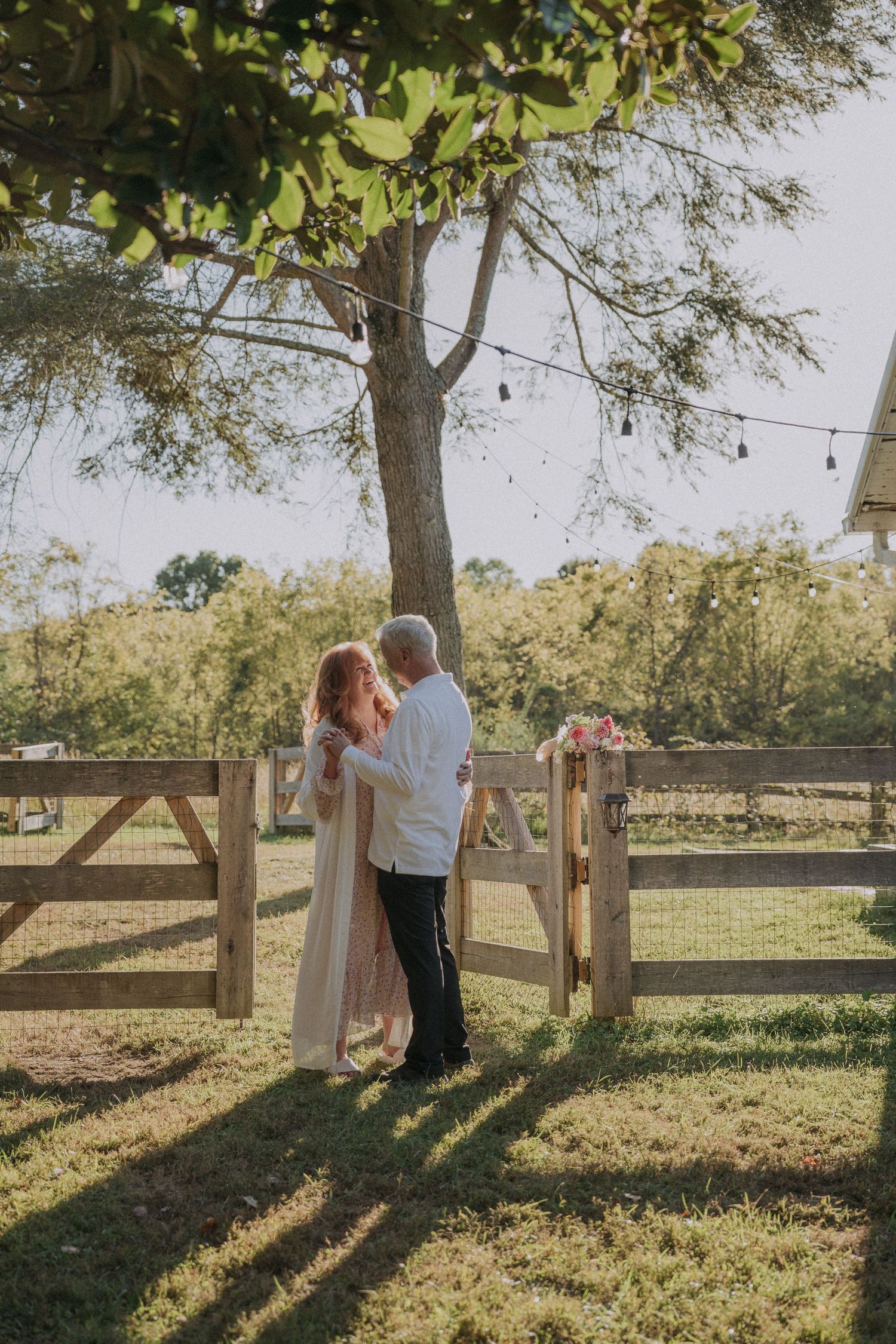Rustic outdoor scene with couple dancing by a wooden fence and string lights at sunset.