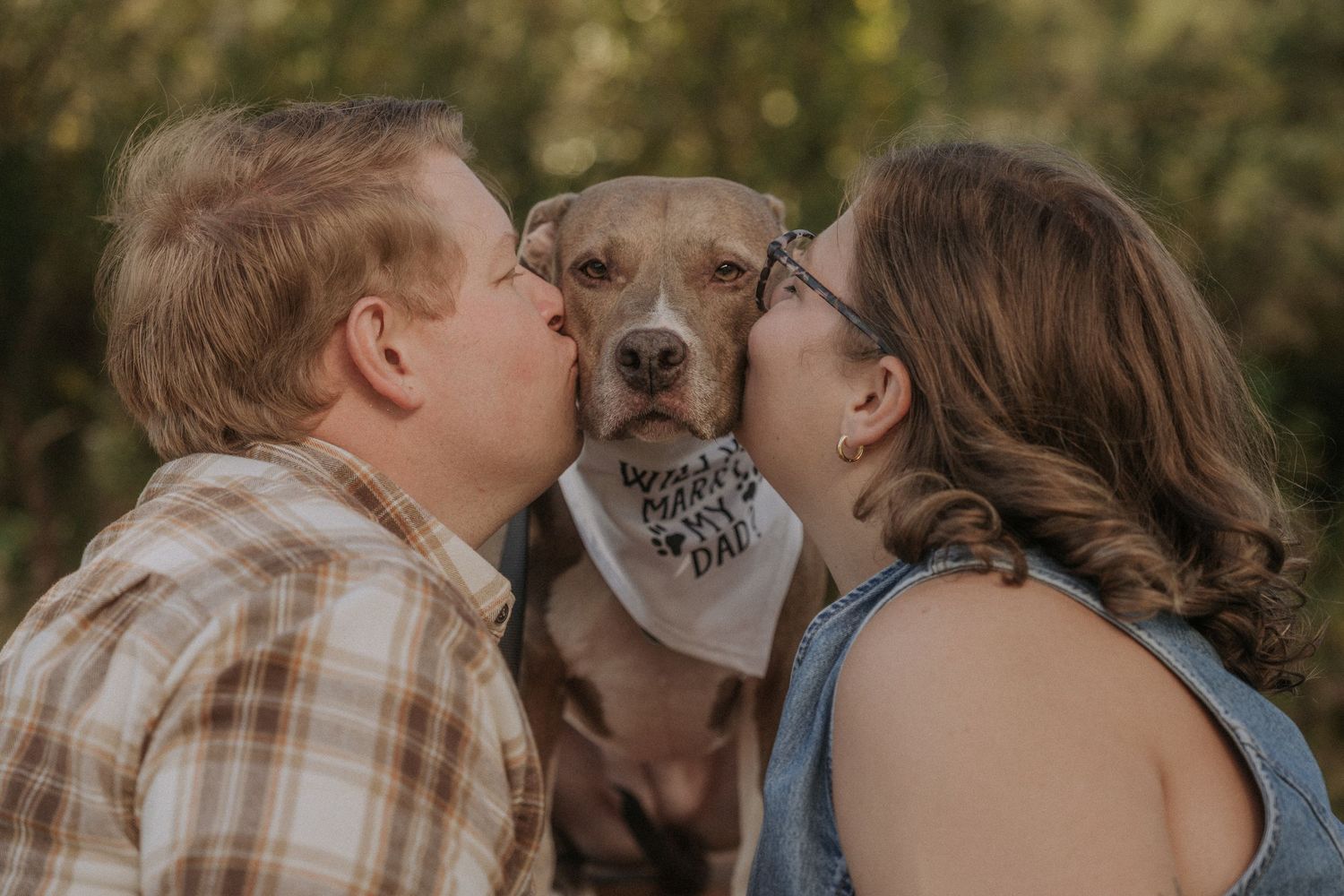 Intimate outdoor portrait of a couple kissing their dog with a tree background.