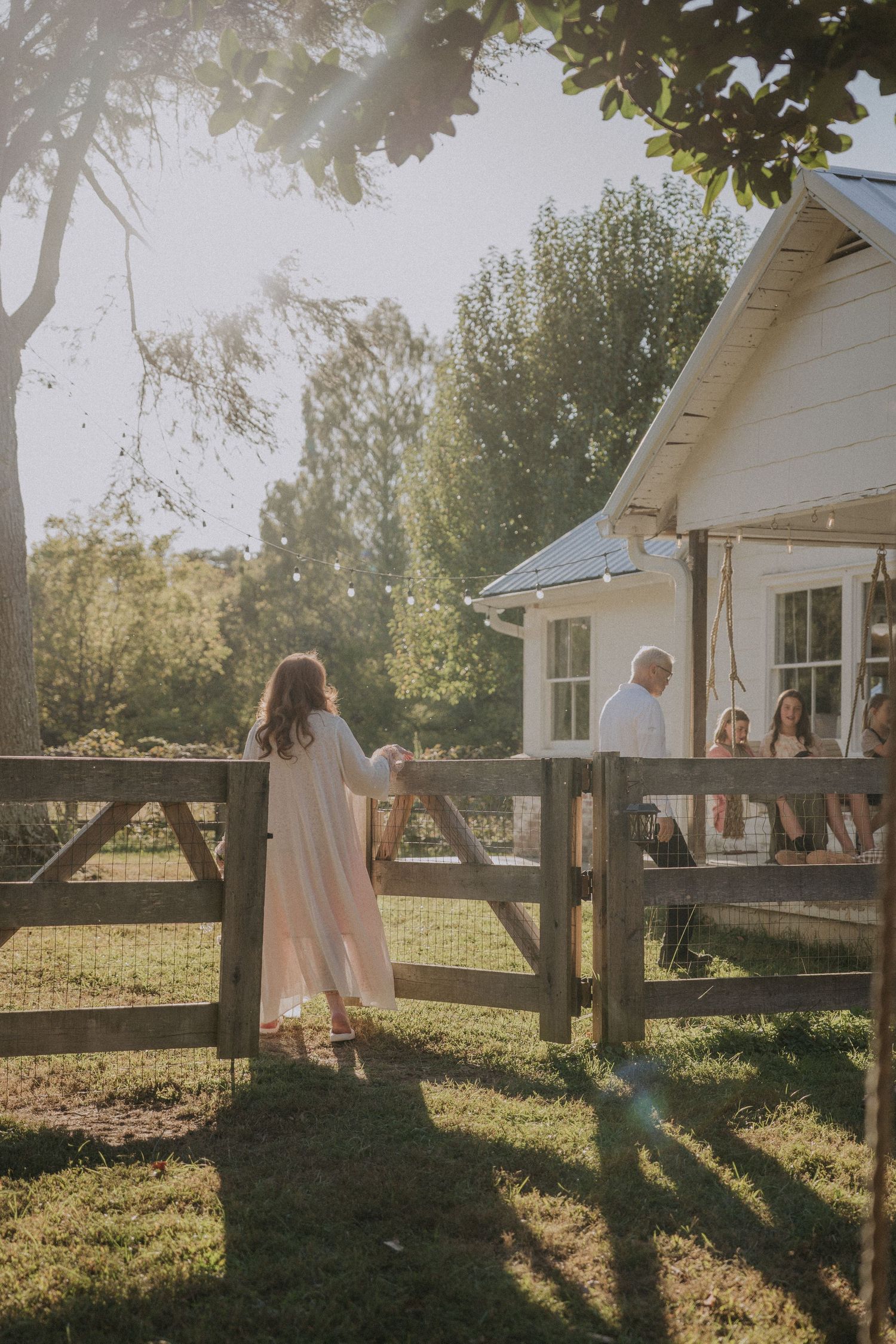 Sun-drenched outdoor scene with white farmhouse and wooden fence casting long shadows across a grassy lawn.