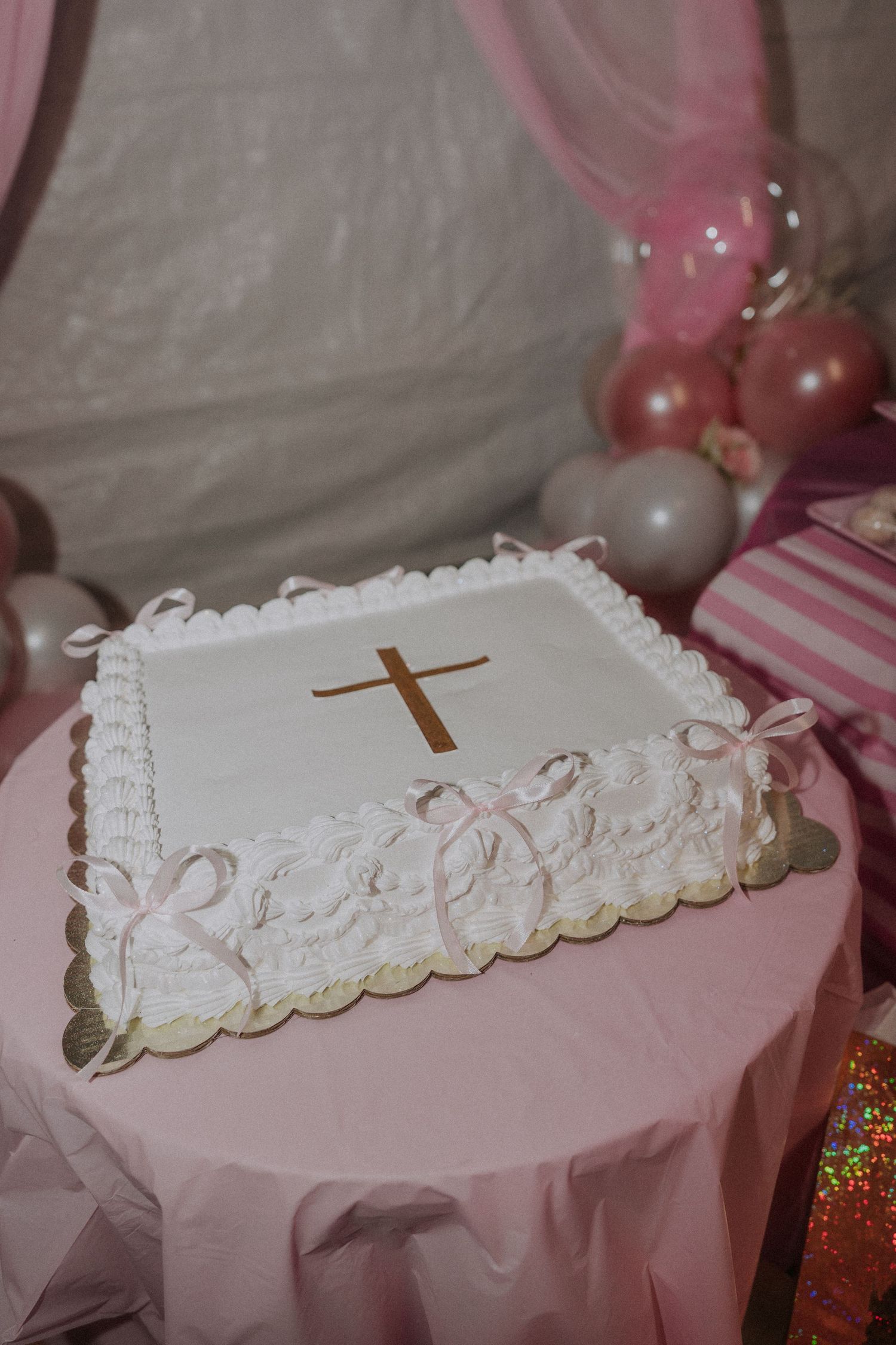White decorated cake with gold cross on pink tablecloth for religious celebration.