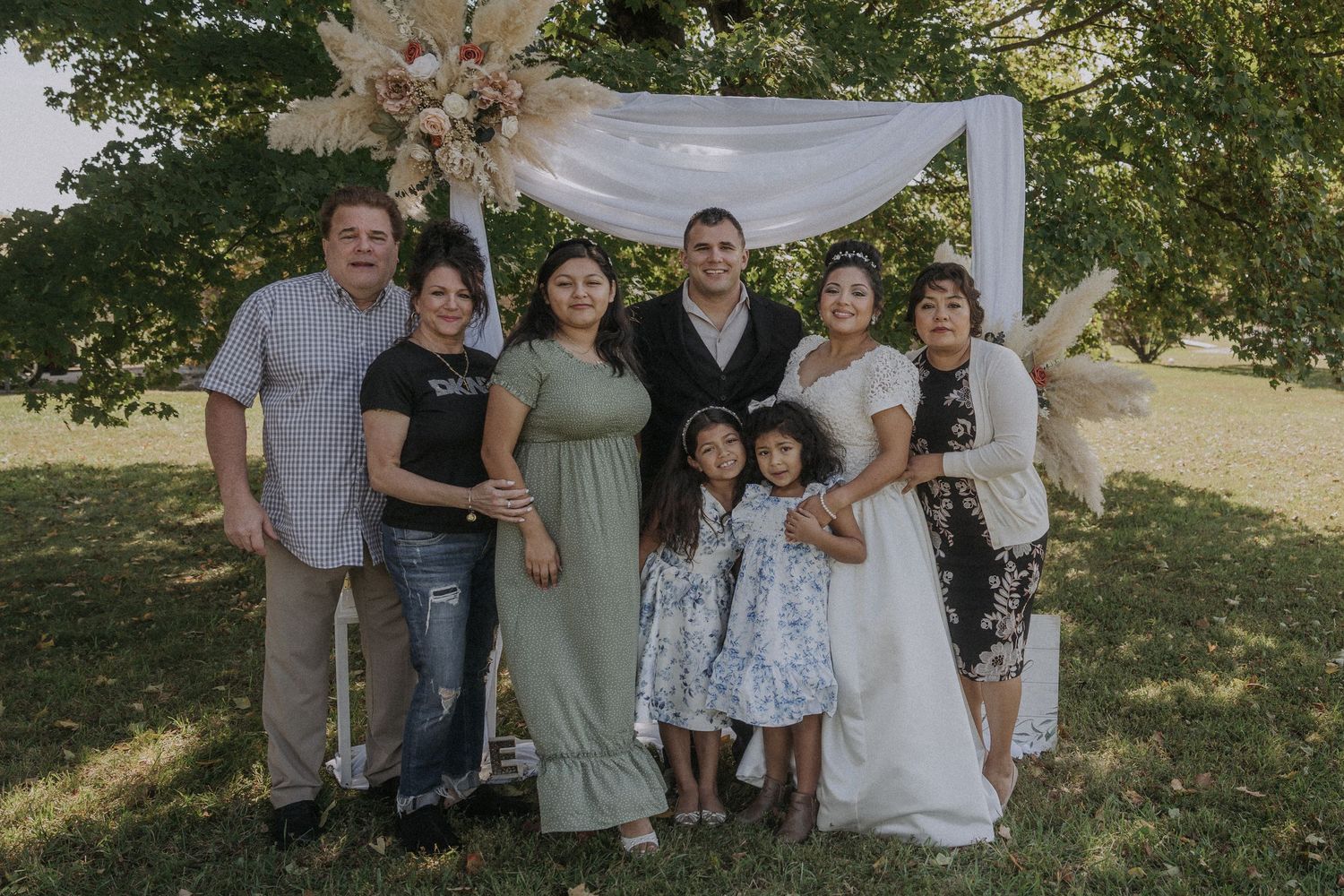 Wedding party and guests gathered for group photo under decorated outdoor arch.