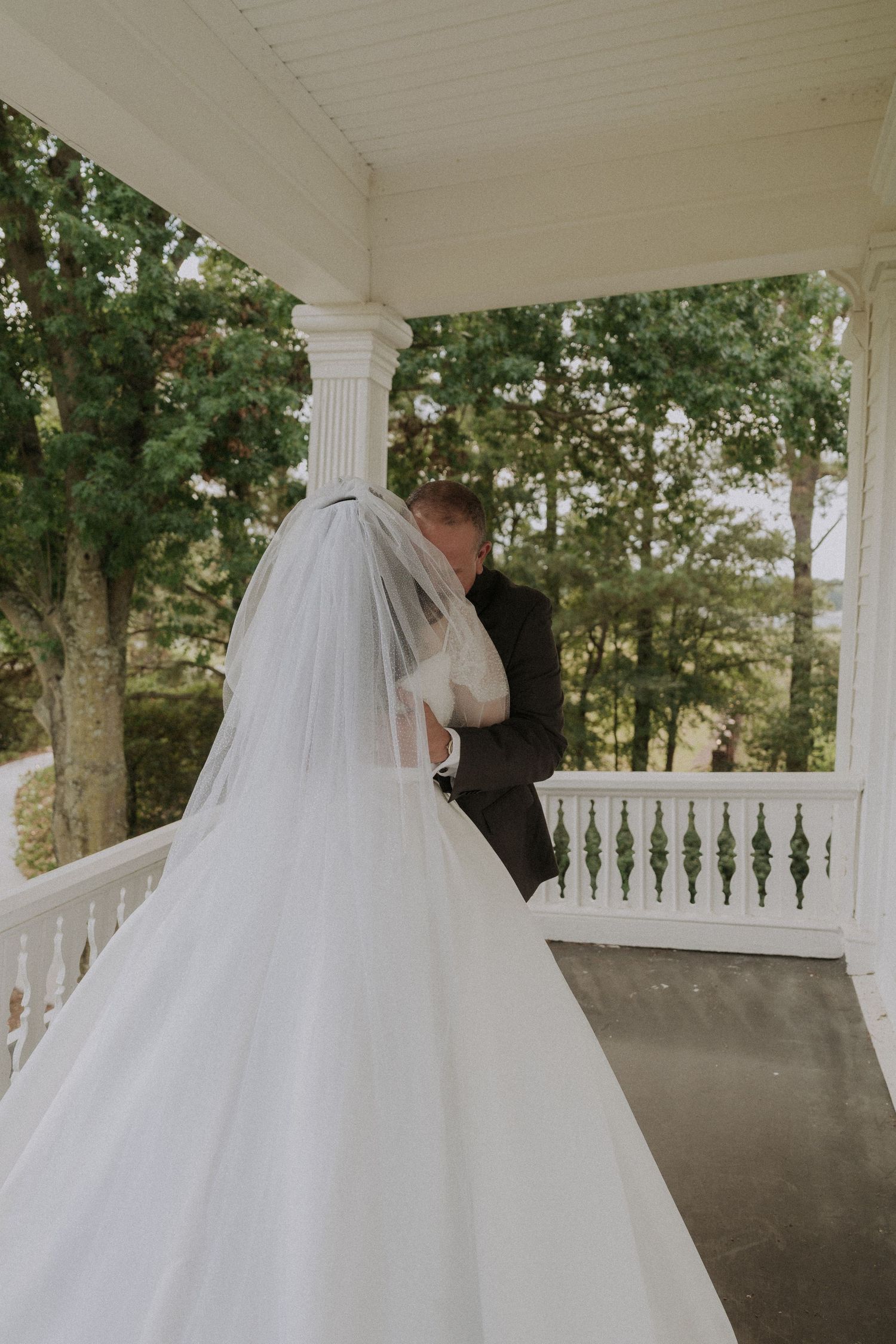 Bride in white gown and veil embracing groom in black suit on elegant white porch with columns.