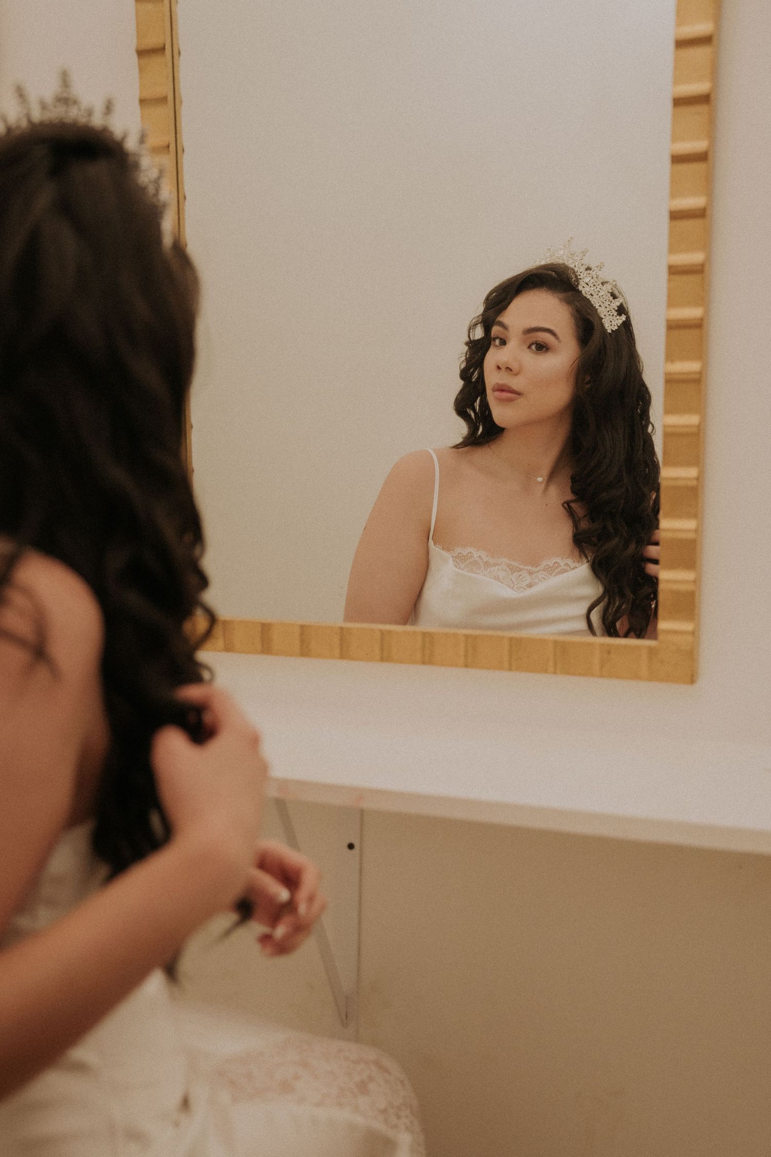 A bride in a white gown and tiara gazes at her reflection in a gold-framed mirror while getting ready.