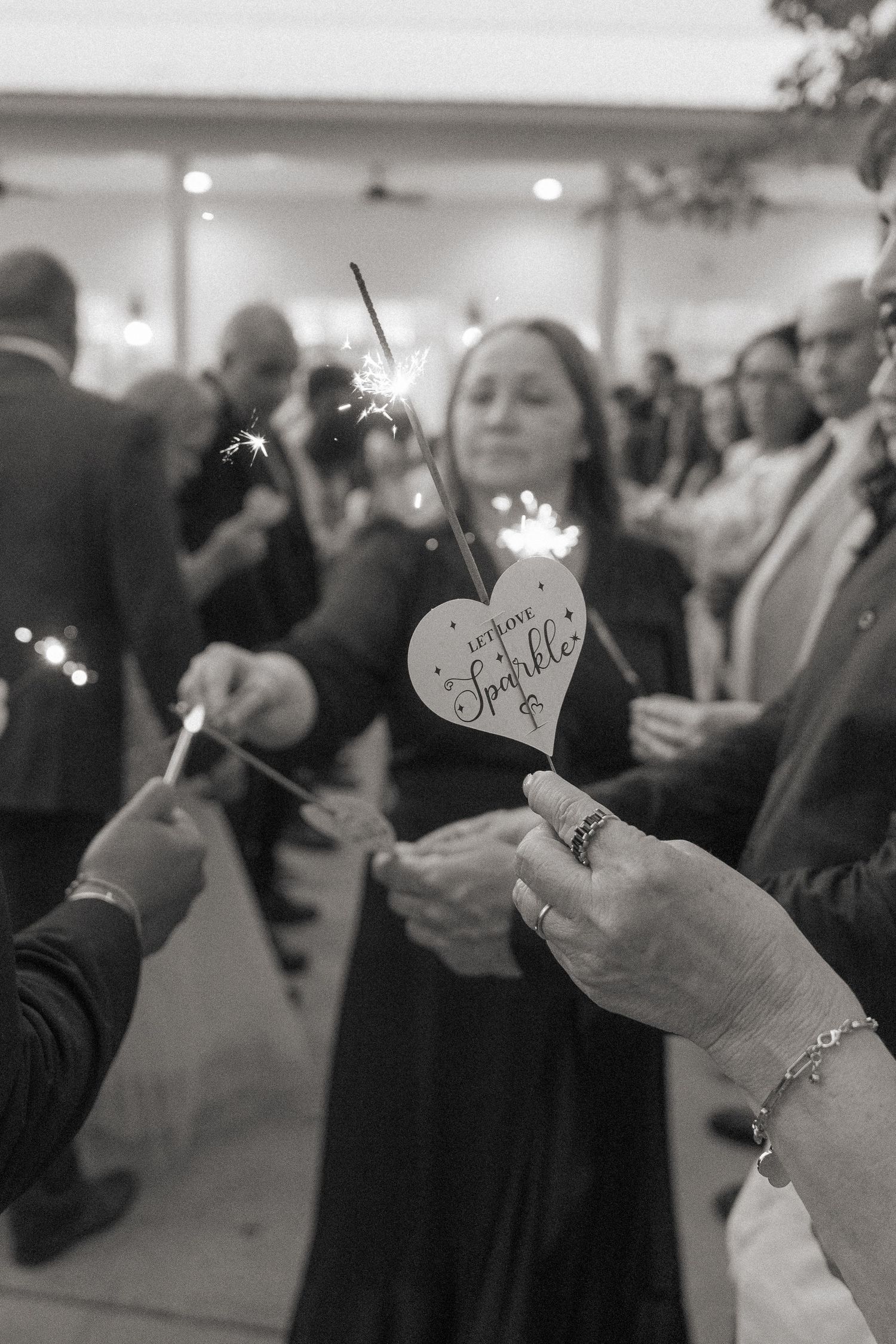Black and white photo of wedding guests holding sparklers and a heart-shaped sign reading 'let love sparkle' at a reception.