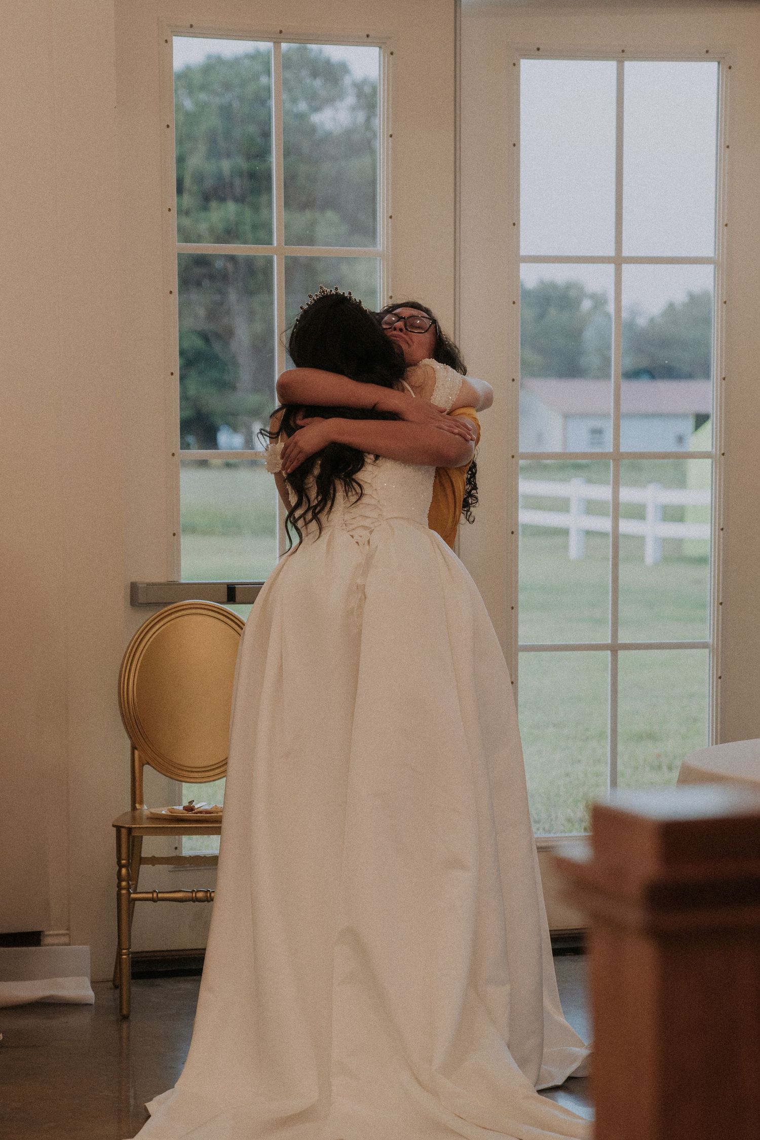 Bride in white ball gown sharing an emotional hug with a guest near large windows at a wedding venue.