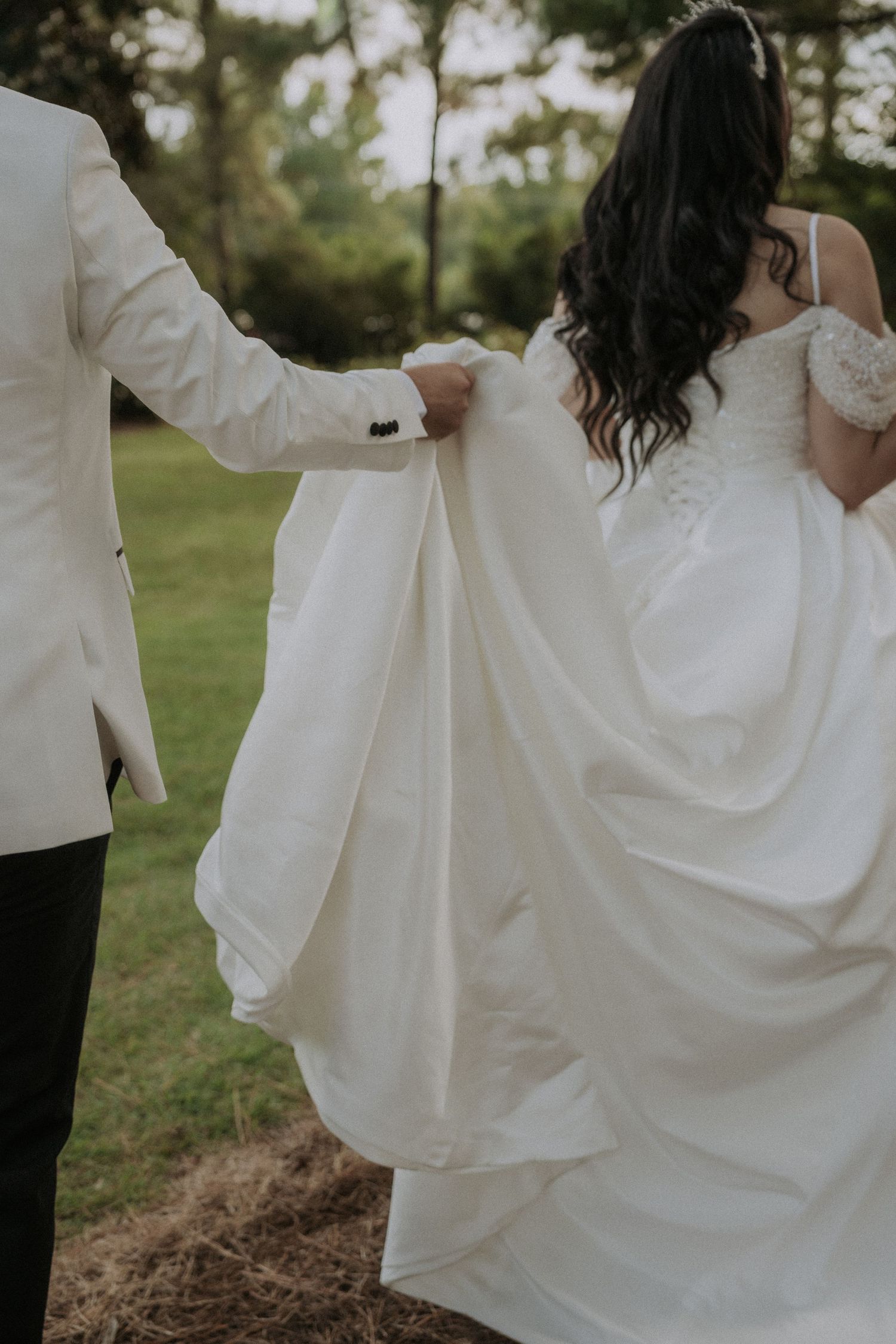 Bride and groom walking outdoors, groom holding white wedding dress train, surrounded by lush green trees.