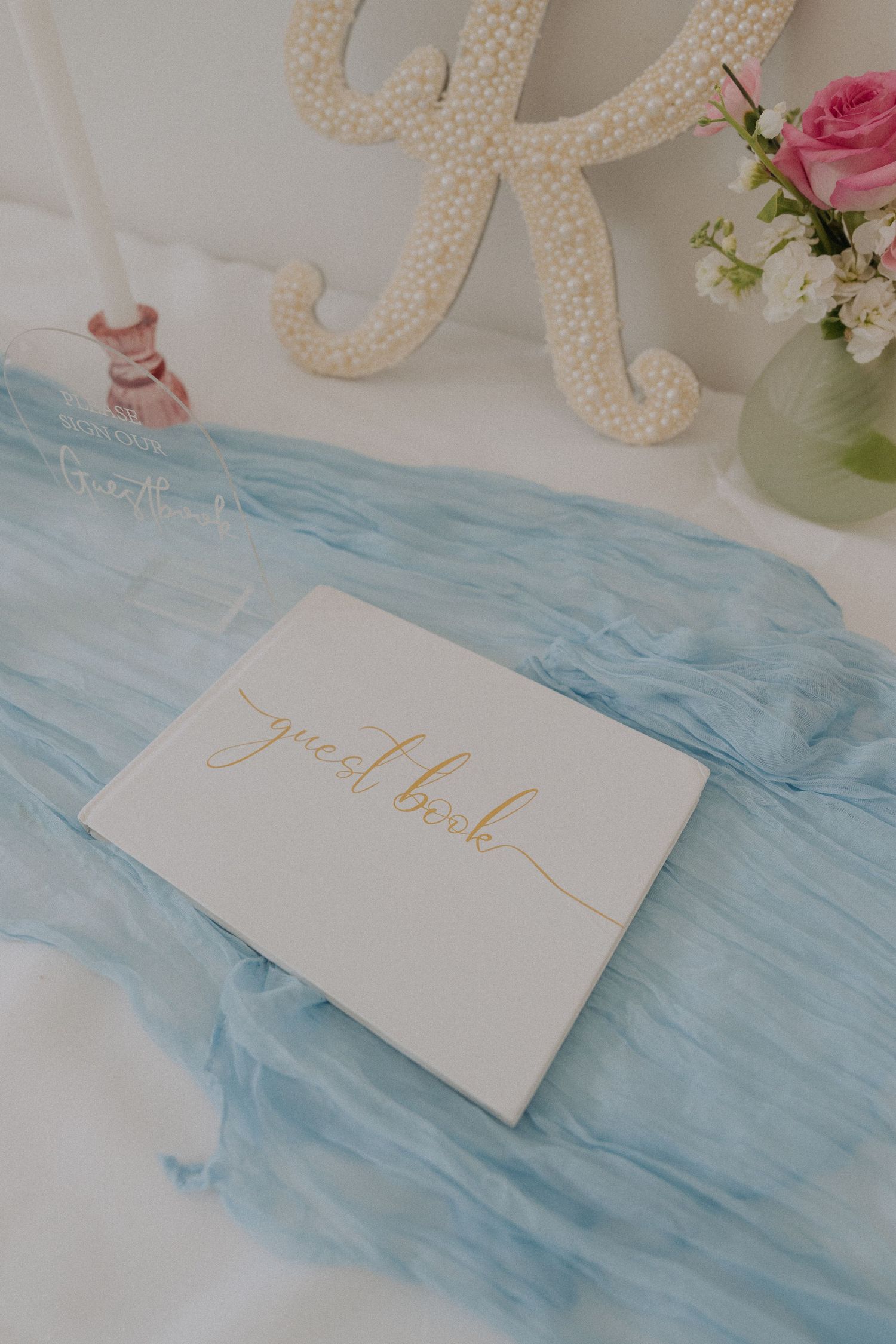 White wedding guest book with gold script on blue tulle fabric beside starfish decor and pink roses.