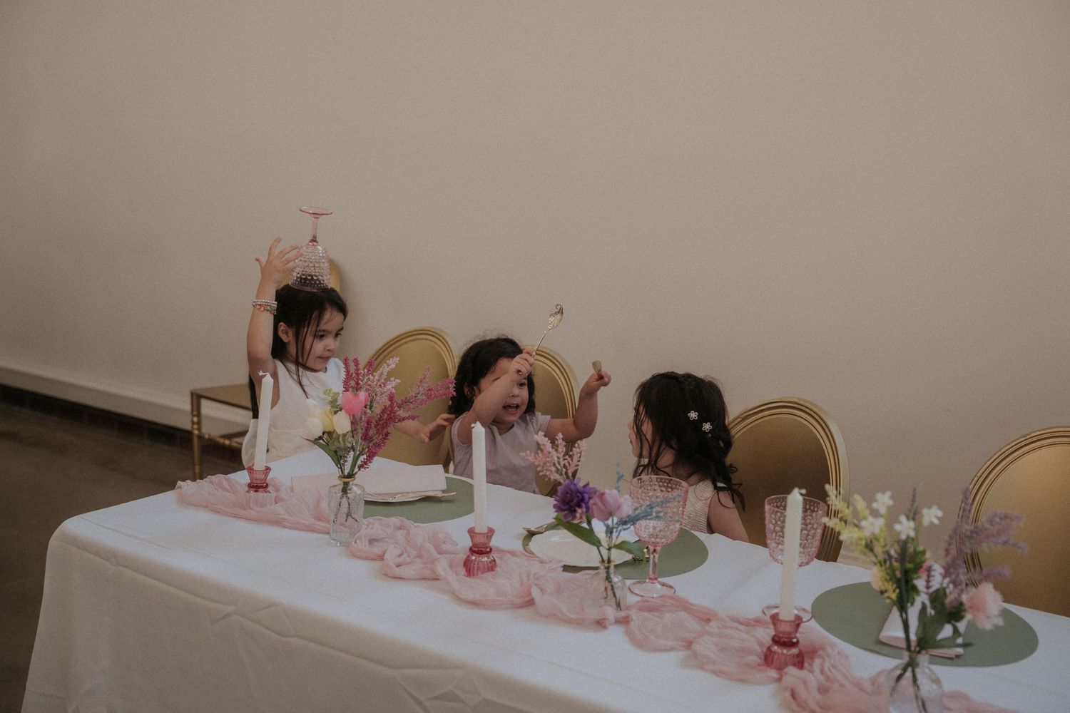 Three young girls sitting at a decorated white table with pink vases, candles, and flowers at a party.