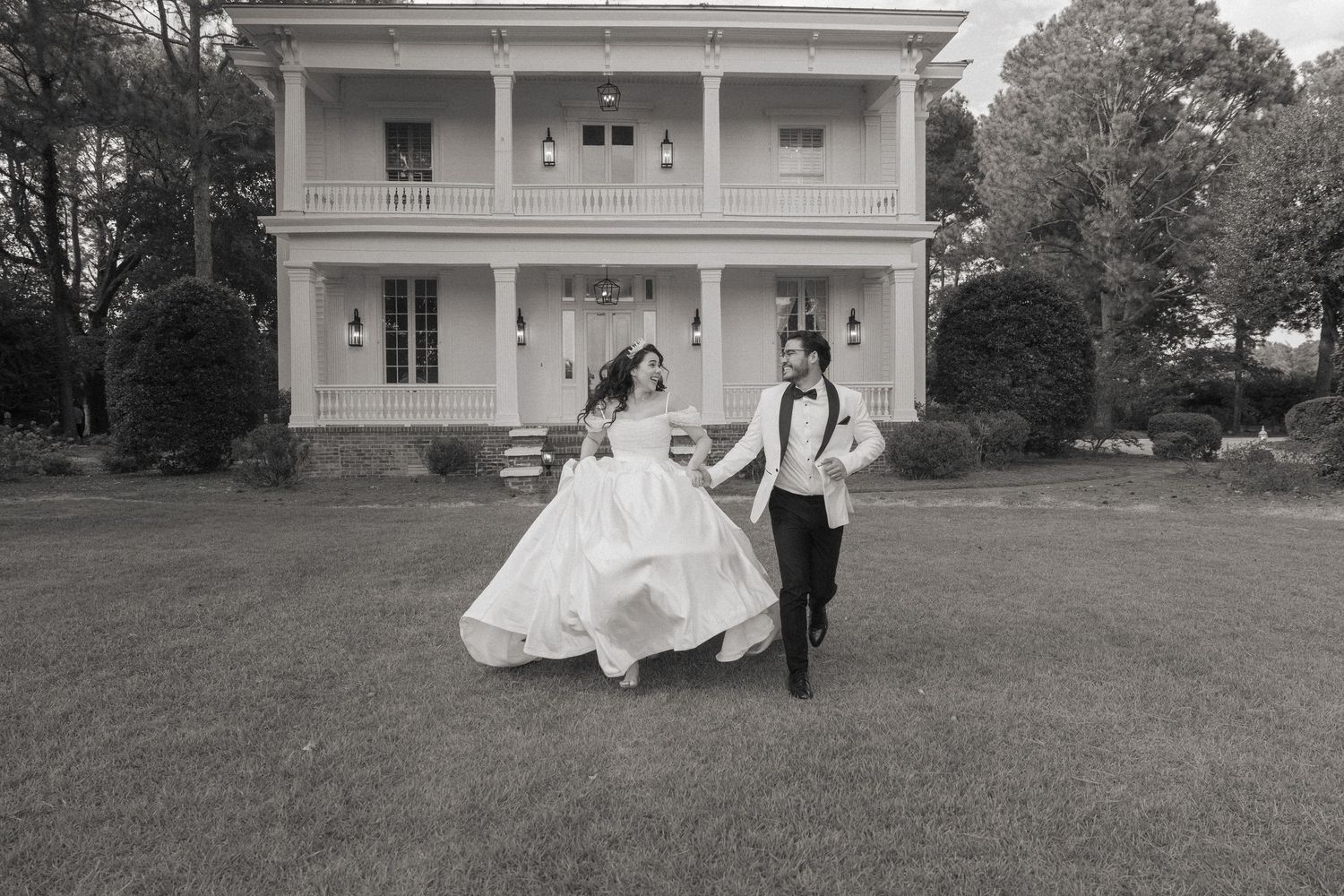 Black and white photo of a bride and groom dancing joyfully on a lawn in front of a grand white Southern mansion.