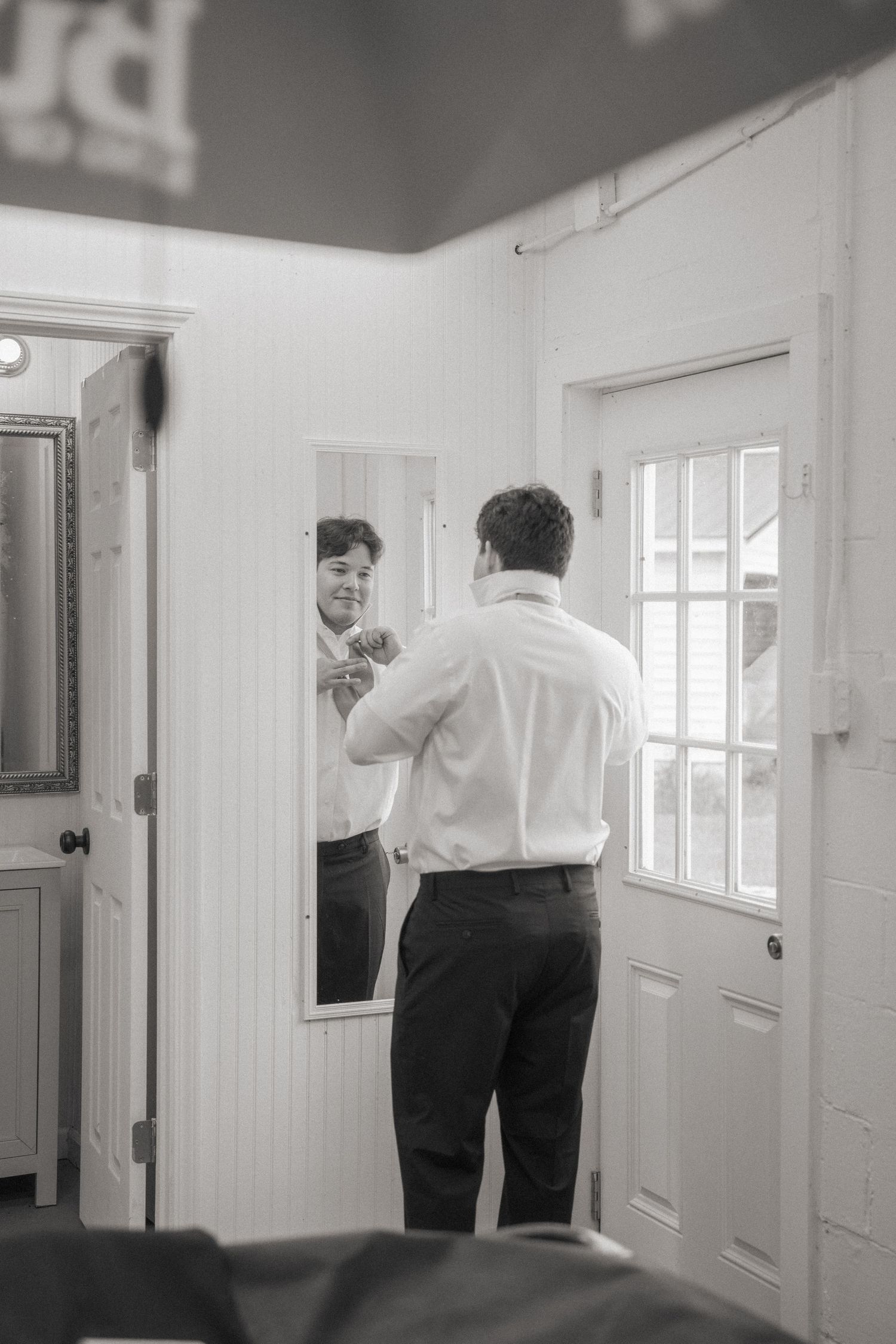 Black and white photo of a groom adjusting his tie in a full-length mirror while getting ready for his wedding.
