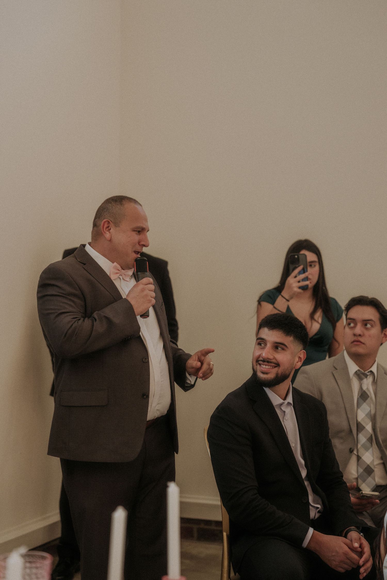 Man in brown suit giving a speech with microphone at a wedding reception while guests smile and record on phone.