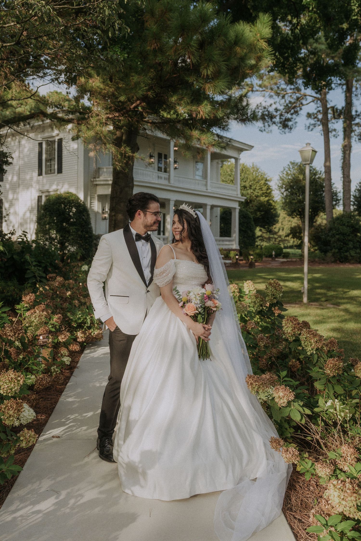 Bride and groom walking together on a garden path outside an elegant white manor on their wedding day.