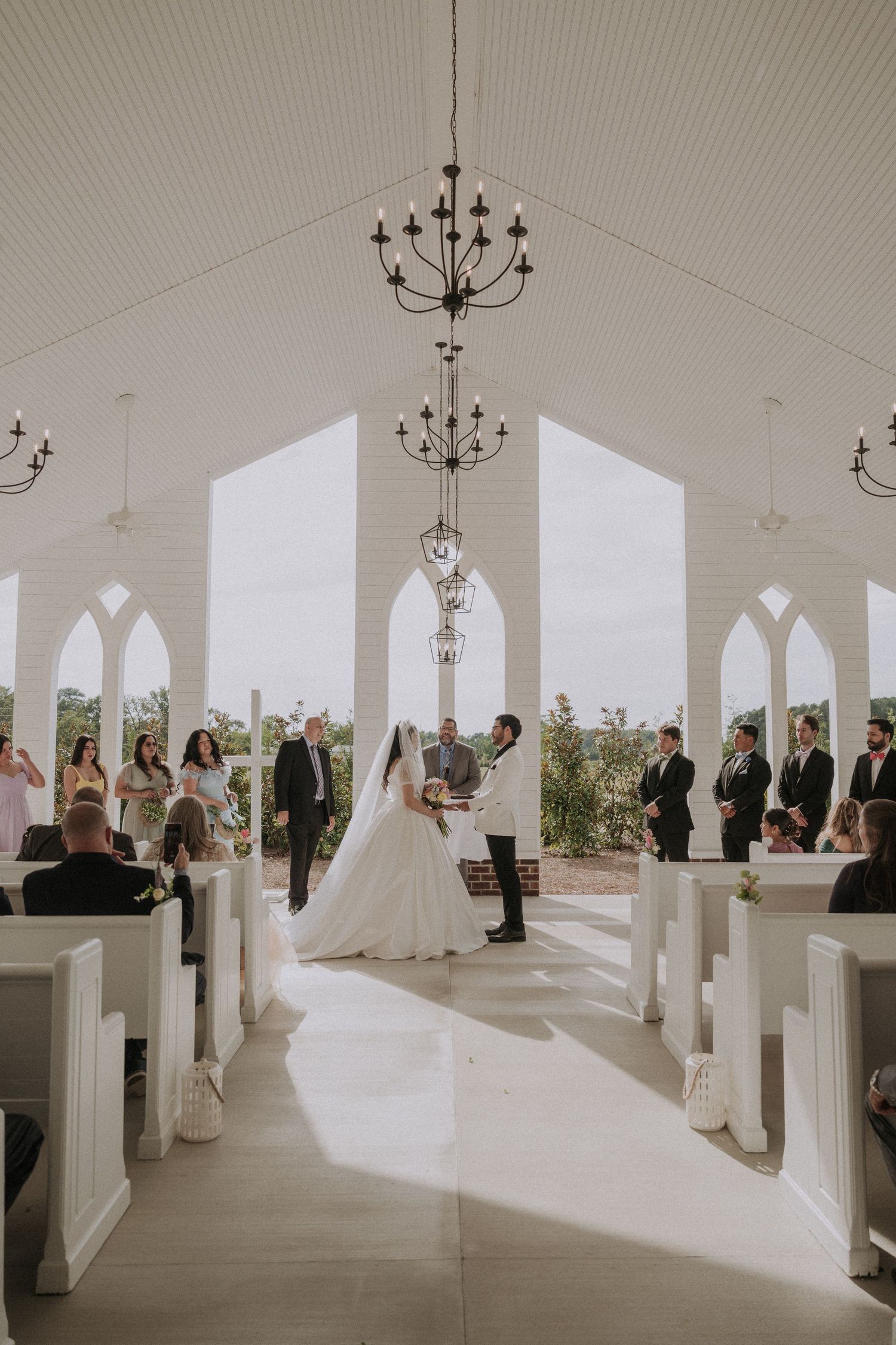 Bride and groom exchange vows at the altar of a stunning white chapel with arched windows and chandelier lighting.