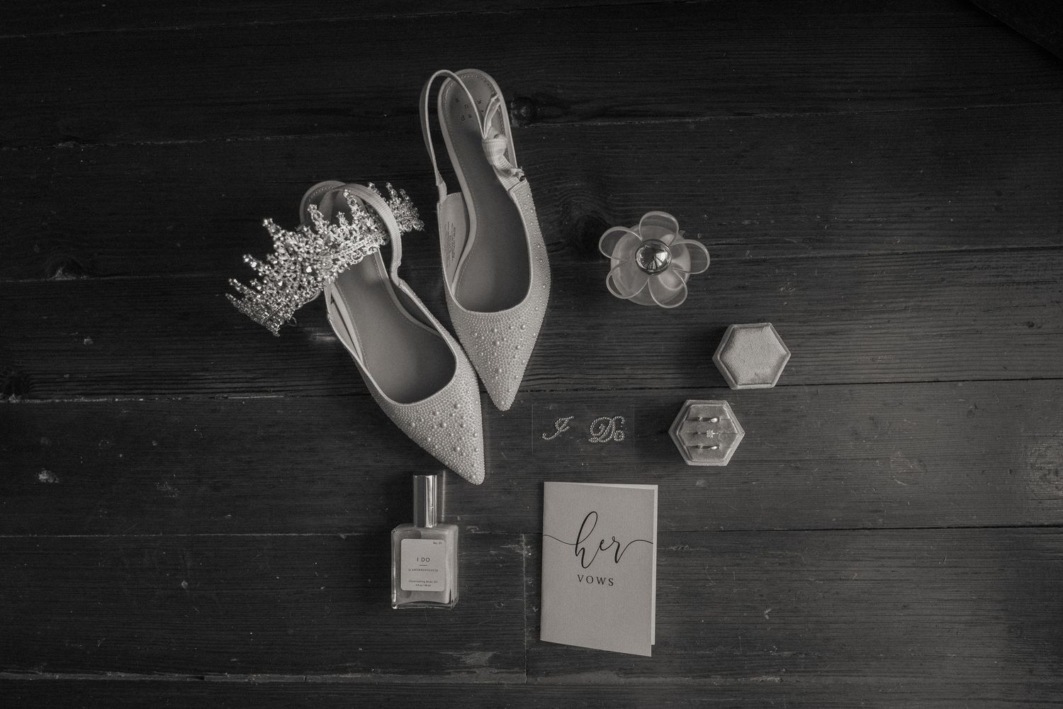 Black and white flat lay of bridal accessories including pointed flats, tiara, perfume, rings, and a wedding card.