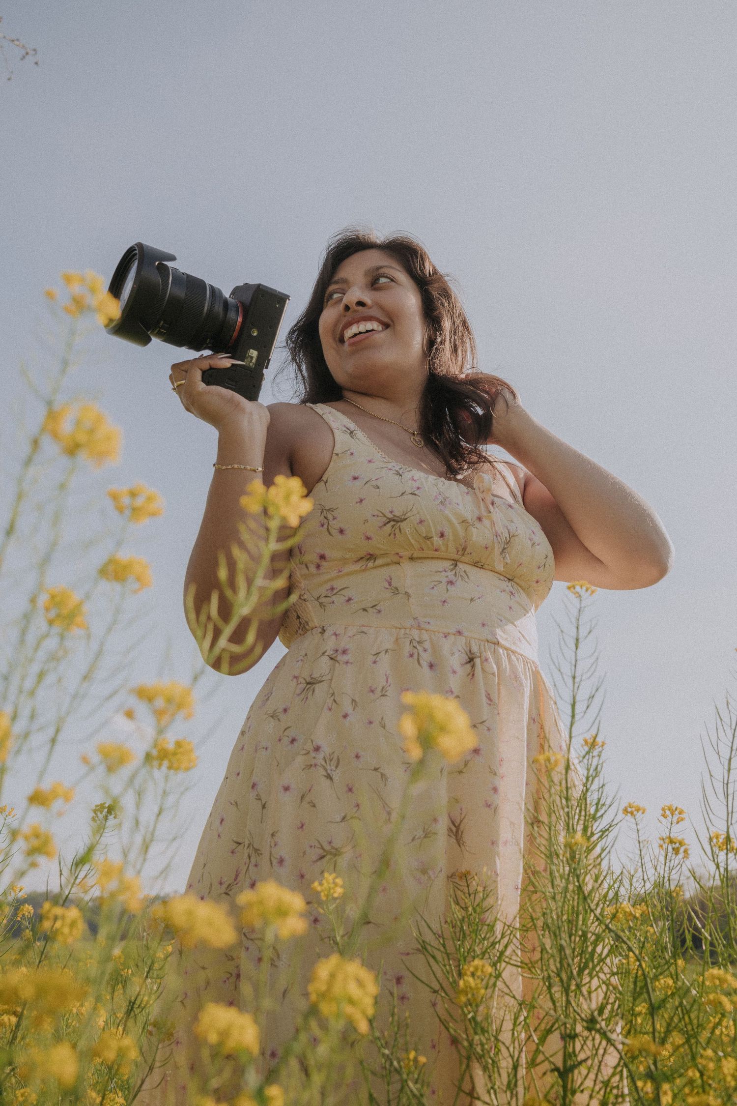 Smiling woman in floral dress holding DSLR camera standing among yellow wildflowers in a sunny field.