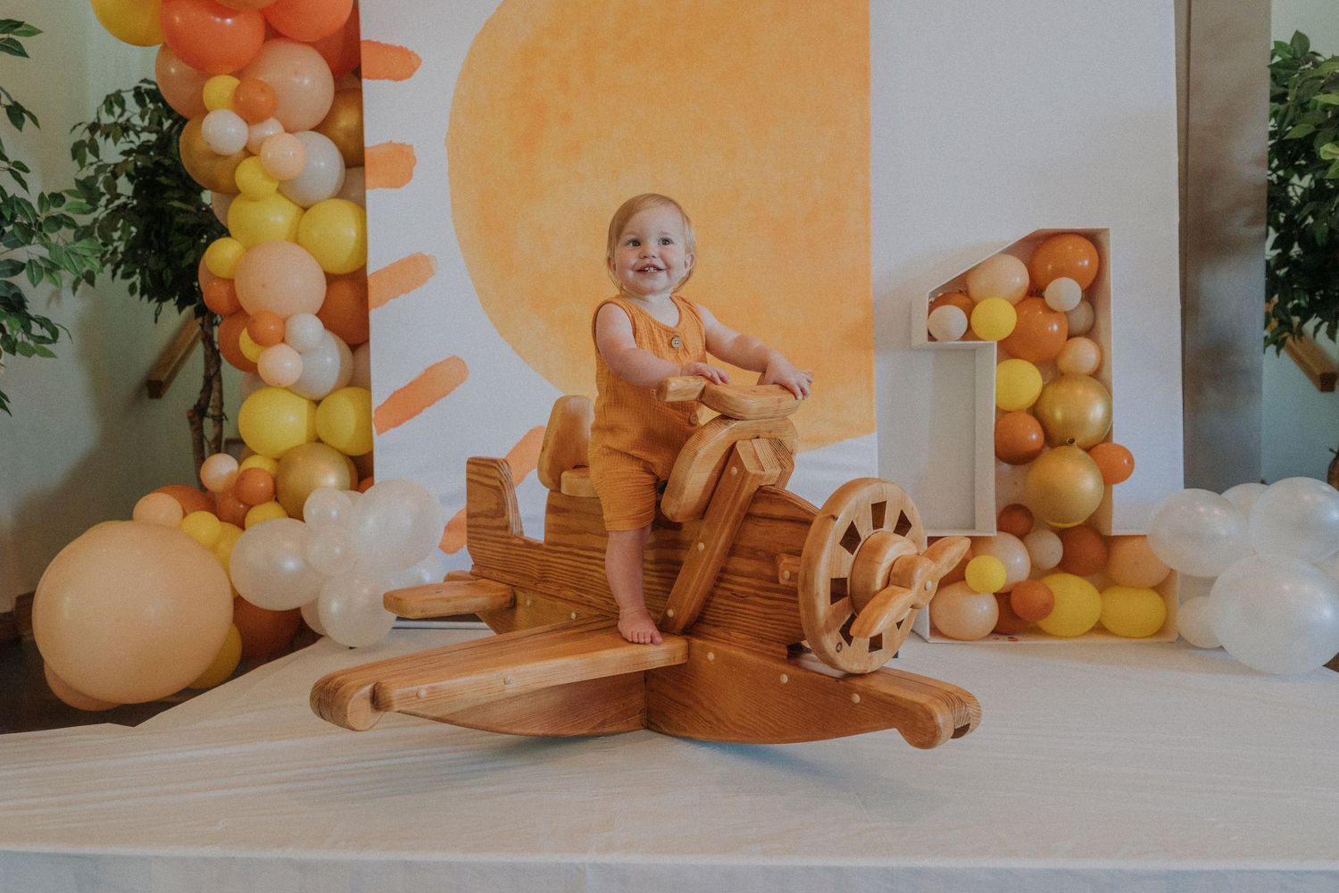 Toddler sitting on wooden rocking horse at first birthday party with yellow and orange balloon garland backdrop.