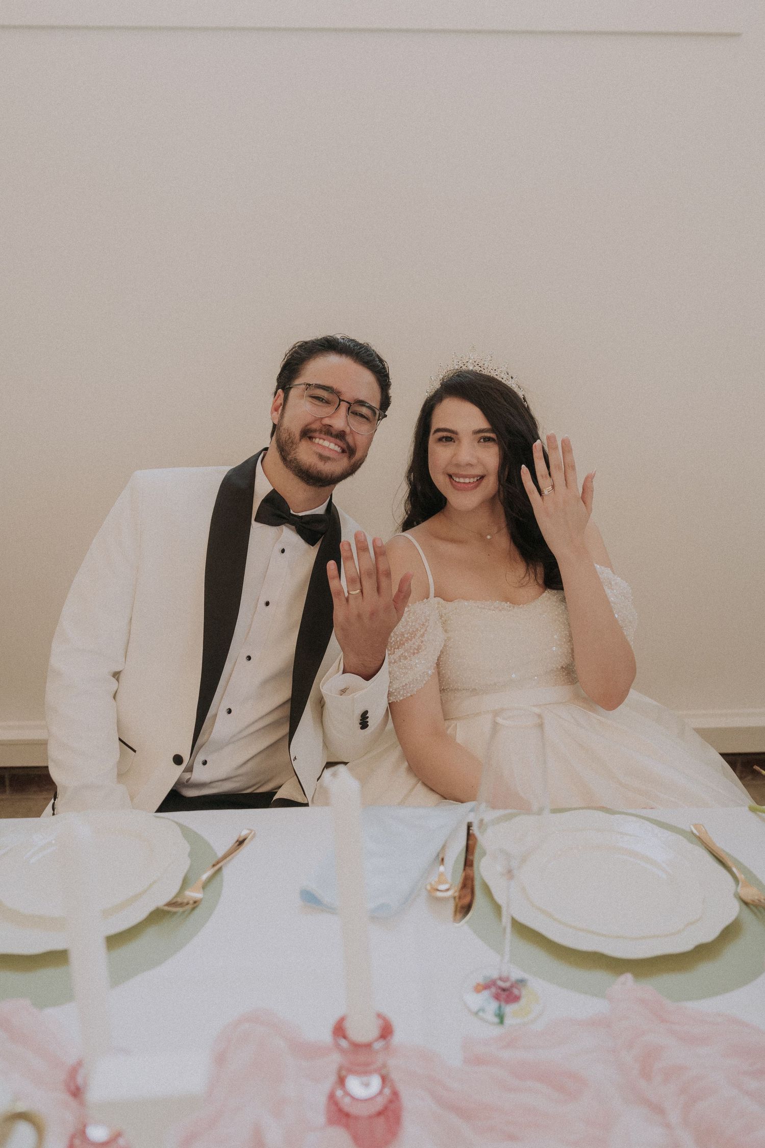 Newlyweds smiling and showing wedding rings at reception table with candles and elegant place settings.