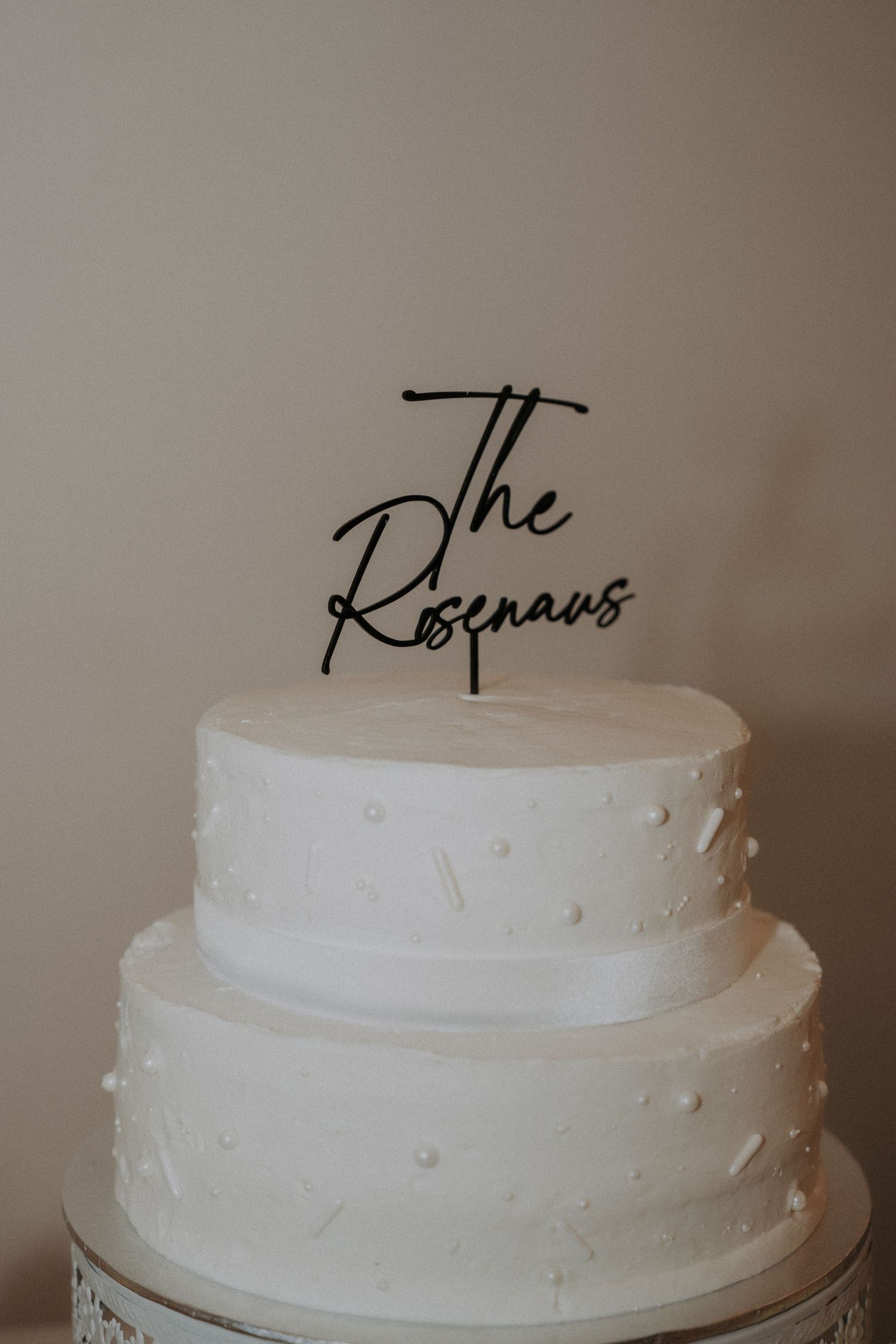 Two-tier white wedding cake with pearl details and black acrylic 'The Rosenaus' cake topper.