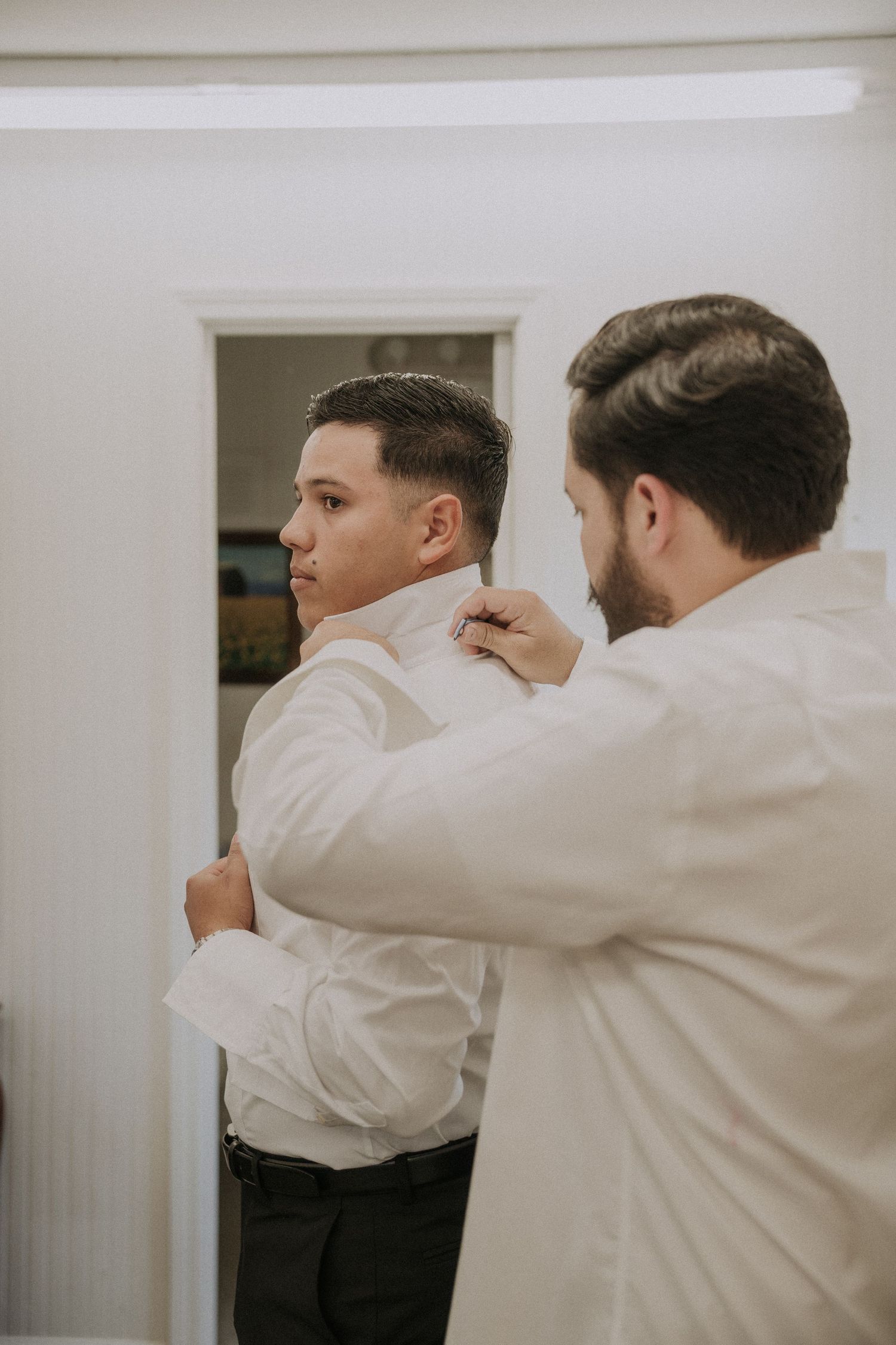 A groomsman helps adjust the collar of another man in a white dress shirt before a wedding ceremony.