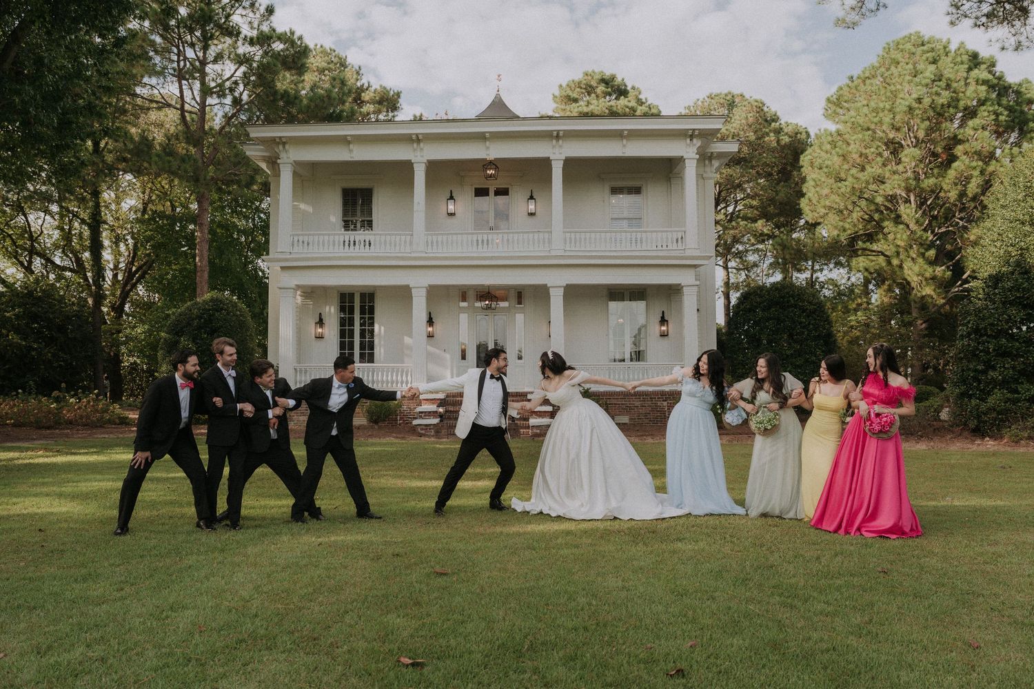 Wedding party posing on lawn before white Southern mansion, groomsmen dancing while bridesmaids in colorful gowns stand nearby.
