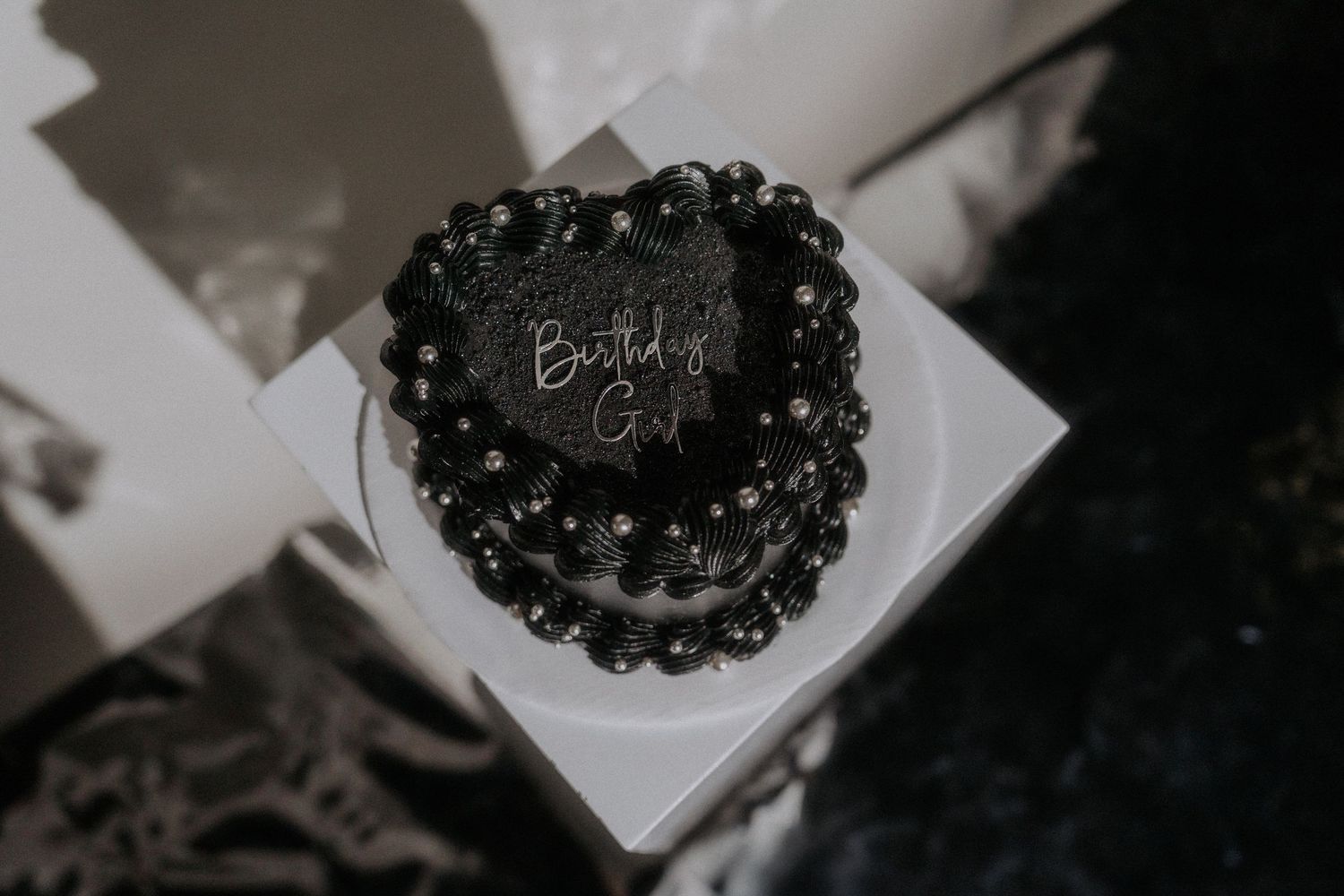 A black vintage heart-shaped birthday cake with dark frosting sits on a white surface in moody lighting.