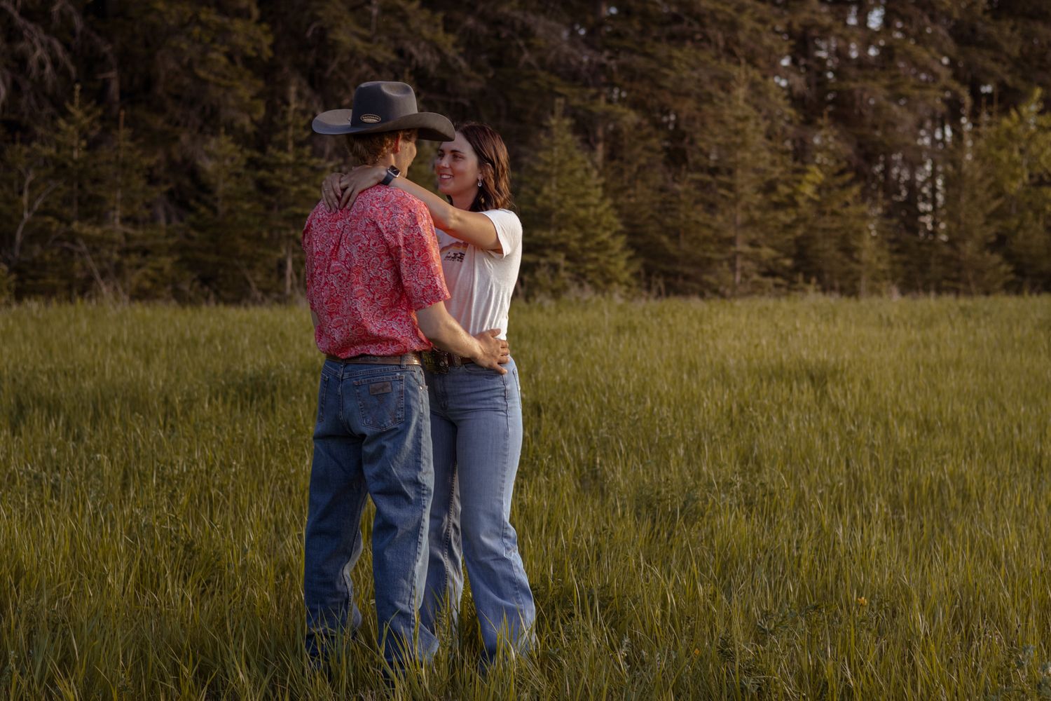 Couple embracing in a lush green meadow with pine forest backdrop, man wearing cowboy hat and red shirt.