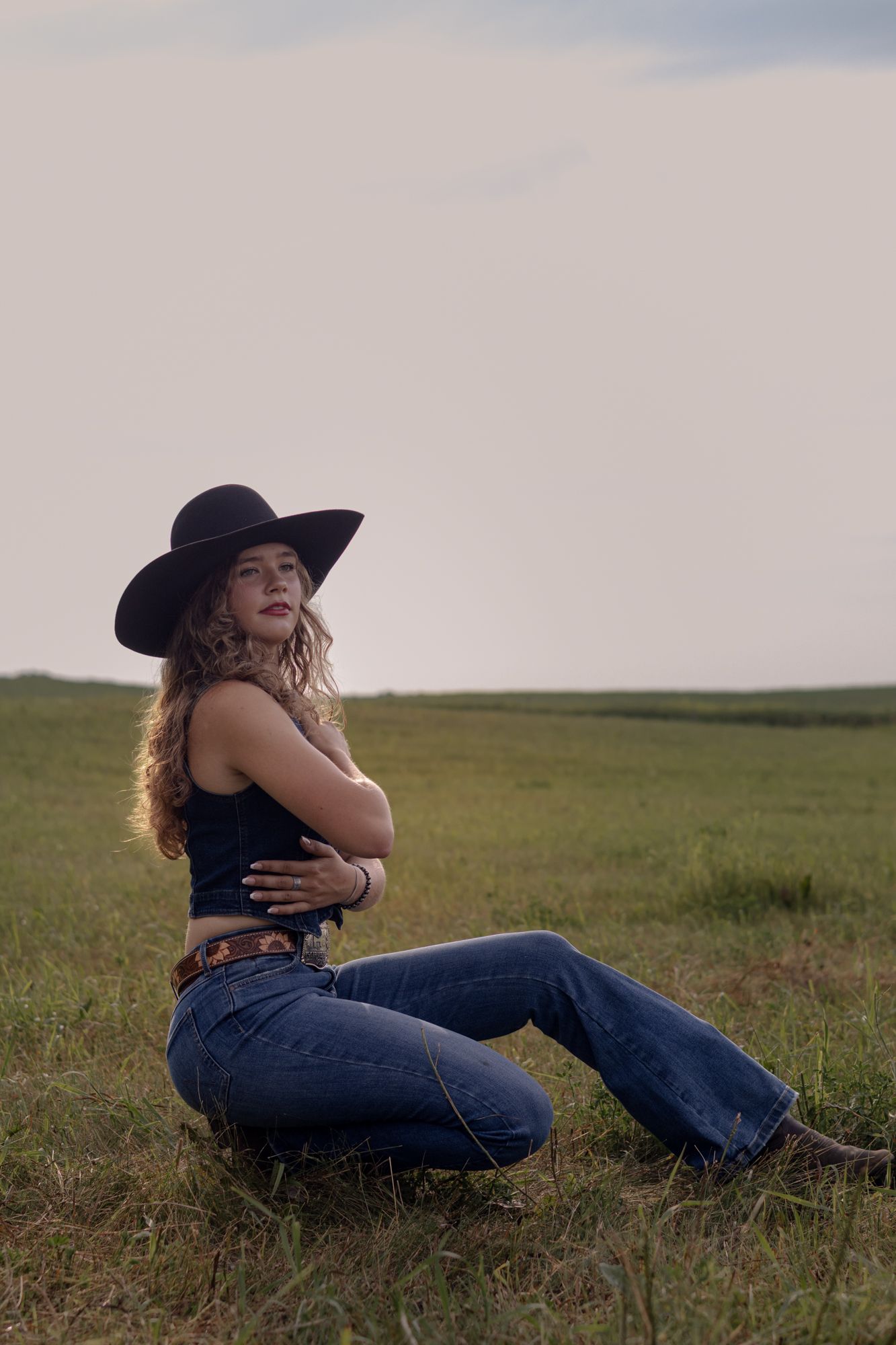 Young woman in black cowboy hat and flare jeans sitting in green field with arms crossed.