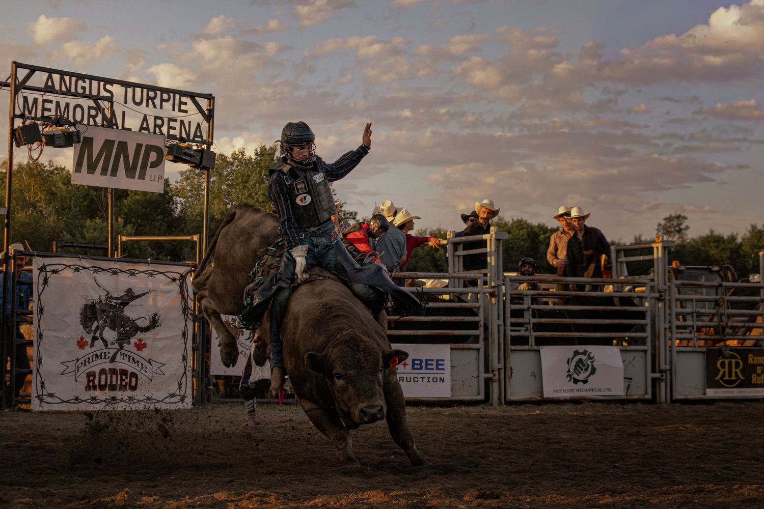 A cowboy rides a bucking bull at Angus Turpie Memorial Arena during a rodeo event at sunset.