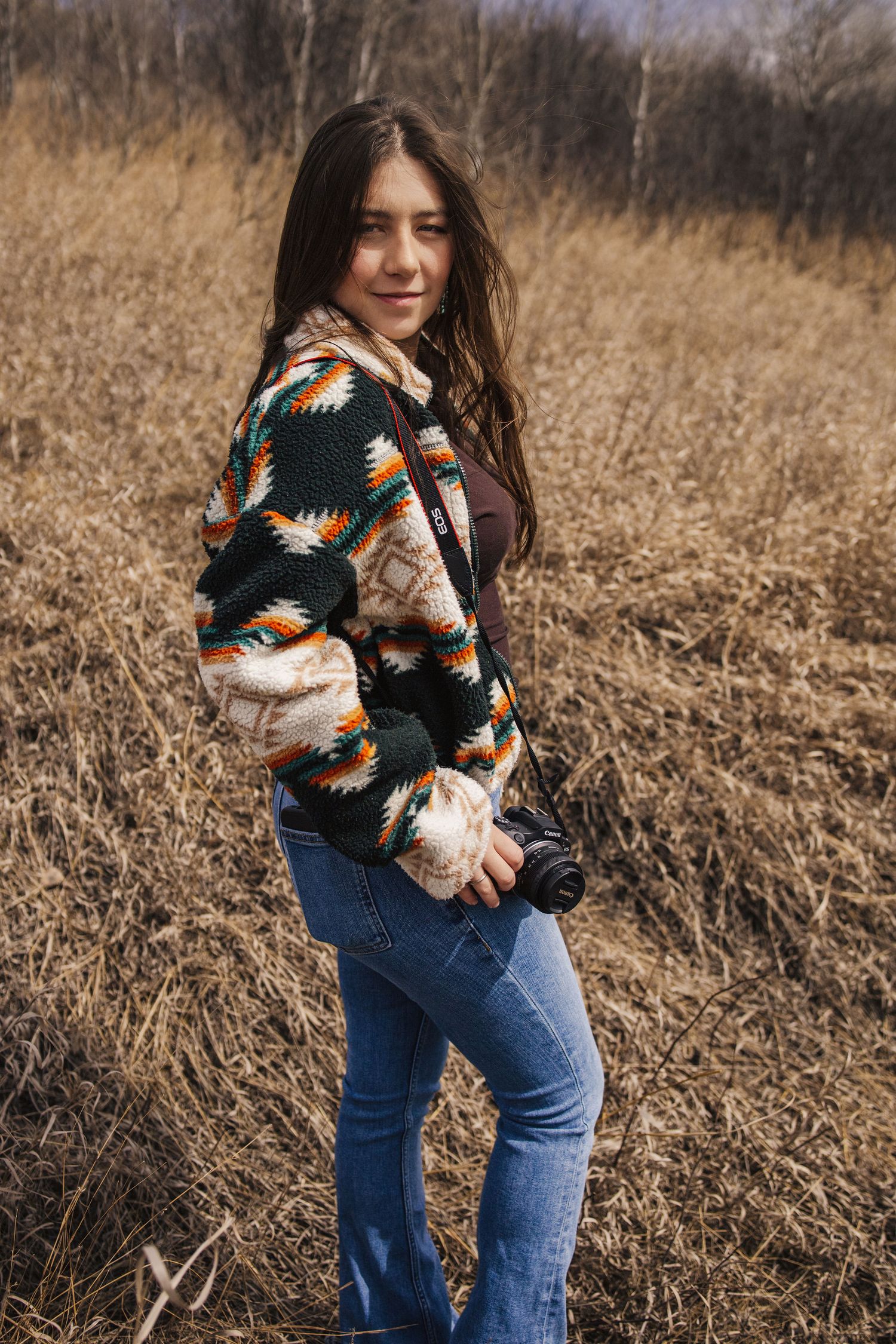 Young woman in patterned jacket holding camera, standing in a golden field of dry grass outdoors.