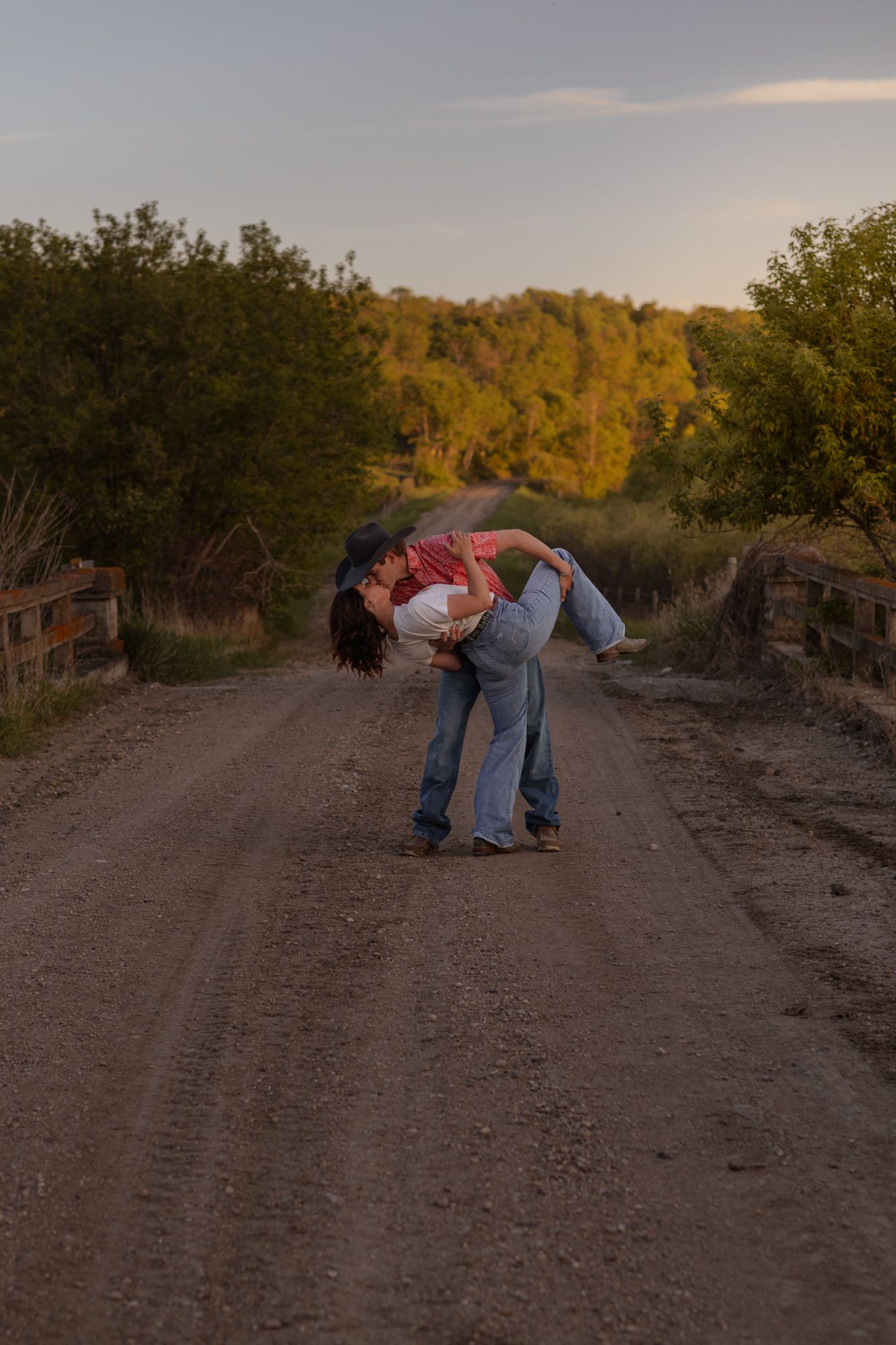 Couple sharing a romantic dip kiss on a rural dirt road at sunset, surrounded by lush green trees.