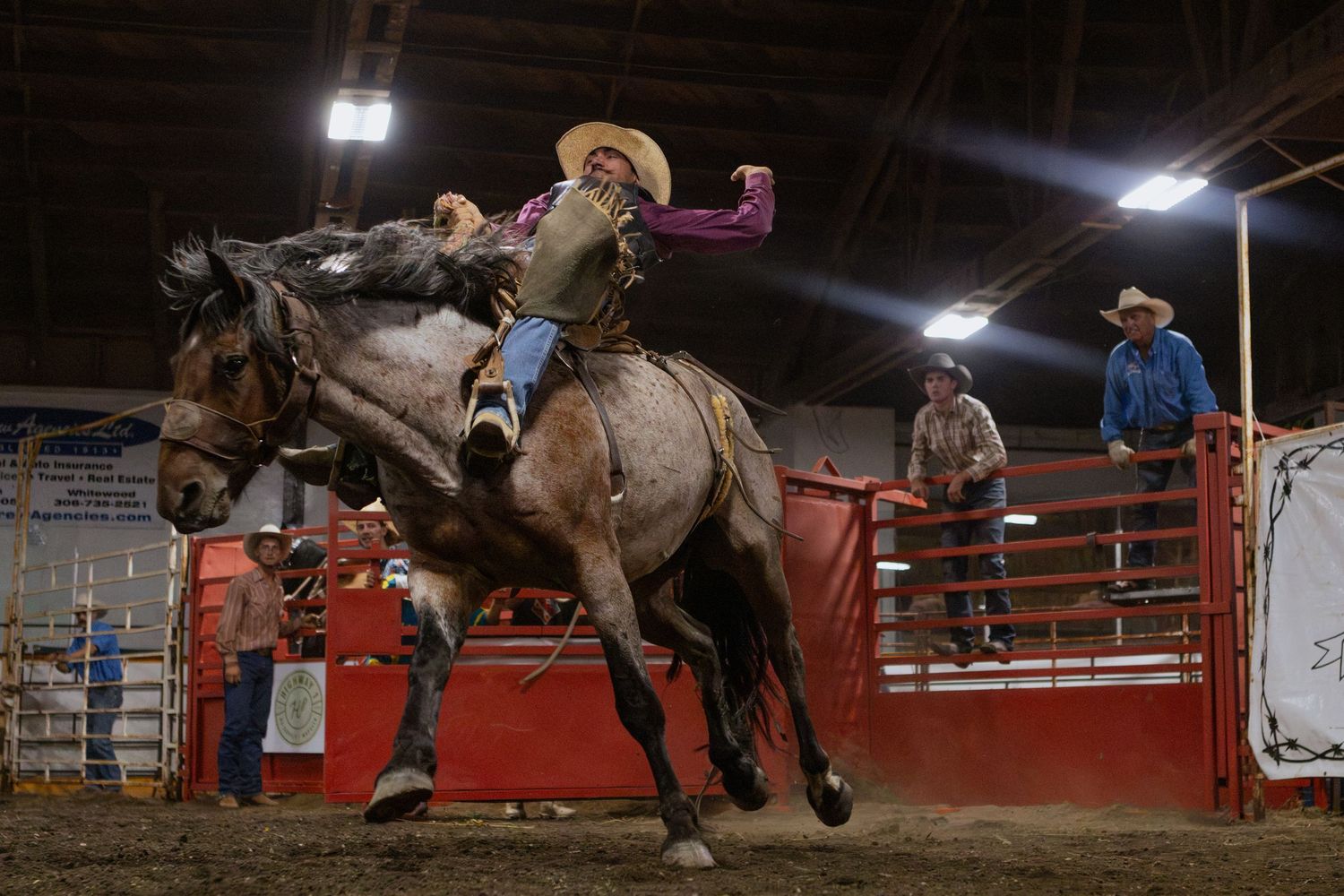 A cowboy rides a bucking bronco at an indoor rodeo arena, with spectators and handlers visible in the background.