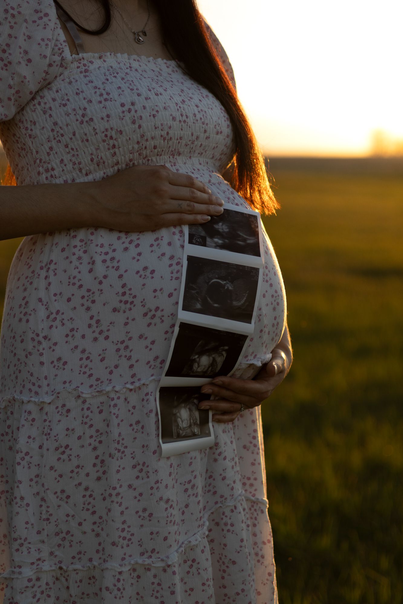 A pregnant woman in a floral dress holds ultrasound images against her belly in a golden field.