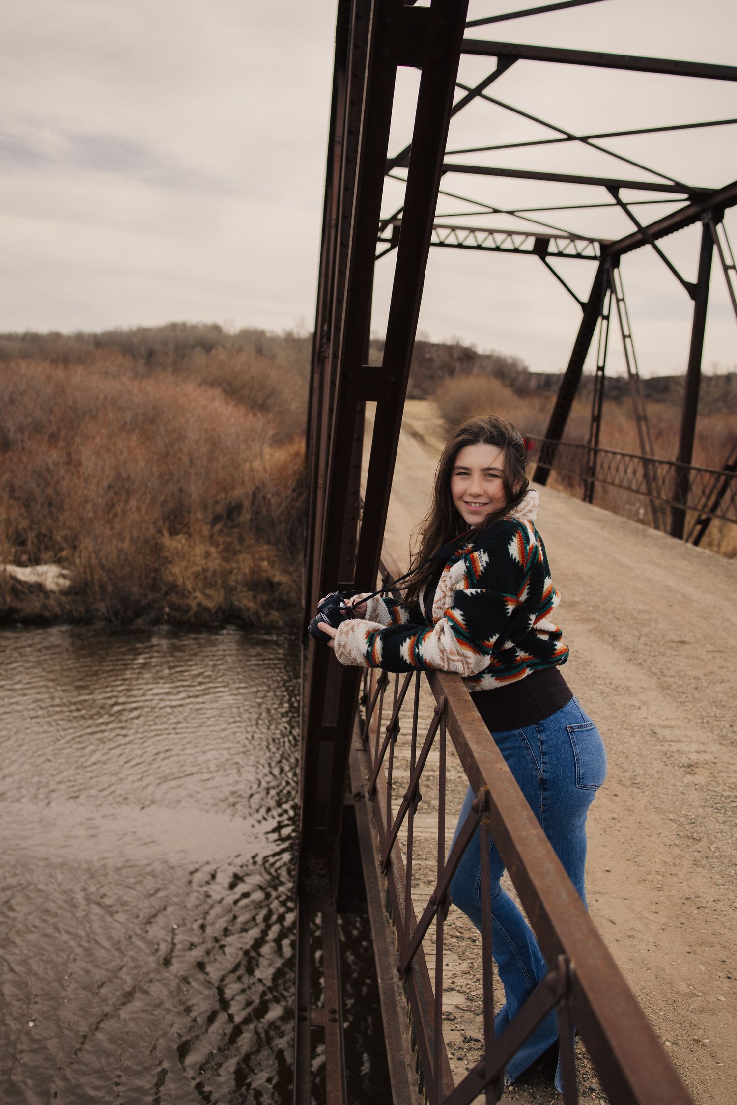 Young woman in colorful sweater and jeans leaning on rustic iron bridge railing over river in autumn.