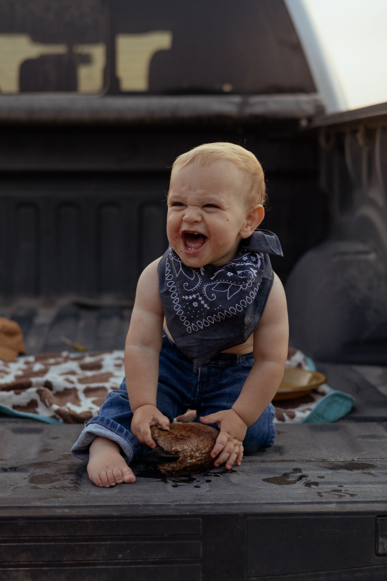 Laughing baby wearing a bandana bib and jeans sits on a truck bed holding a smash cake.