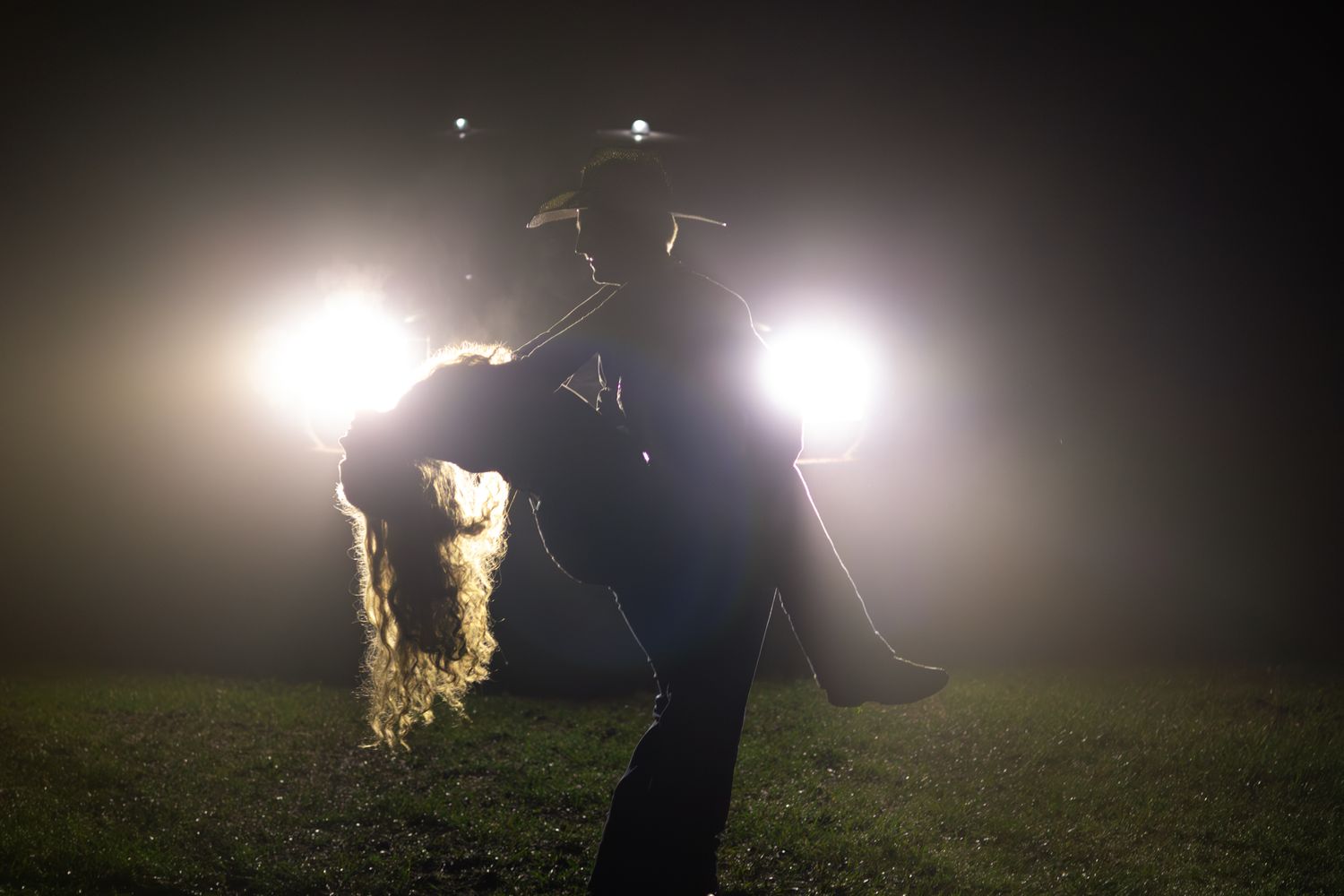 Silhouetted cowboy carrying hay bales in front of bright vehicle headlights at night.