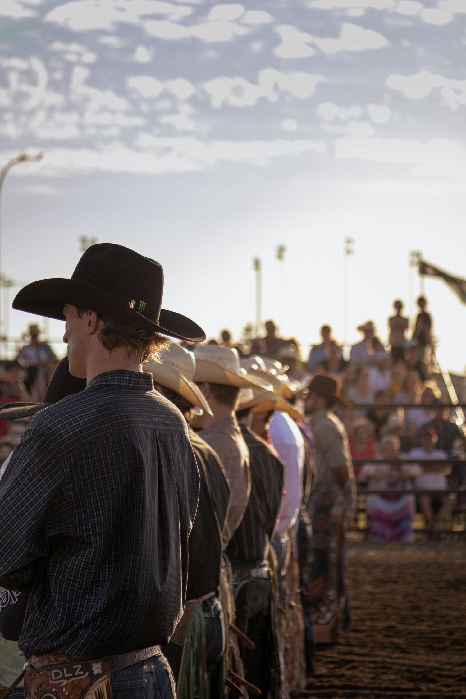 Cowboys in hats line up at a rodeo arena with spectators in the bleachers under a cloudy sky.