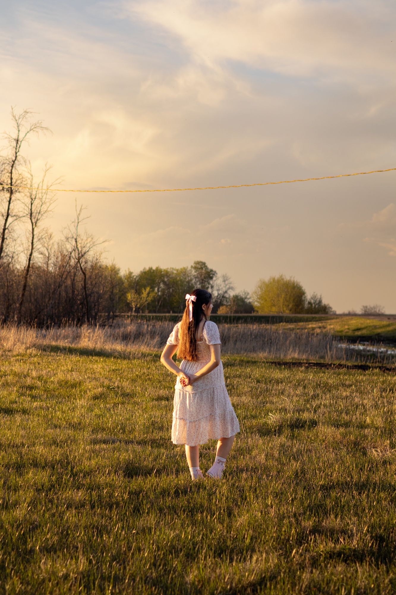 Young girl in white dress standing in golden sunset field, viewed from behind, with trees and cloudy sky.