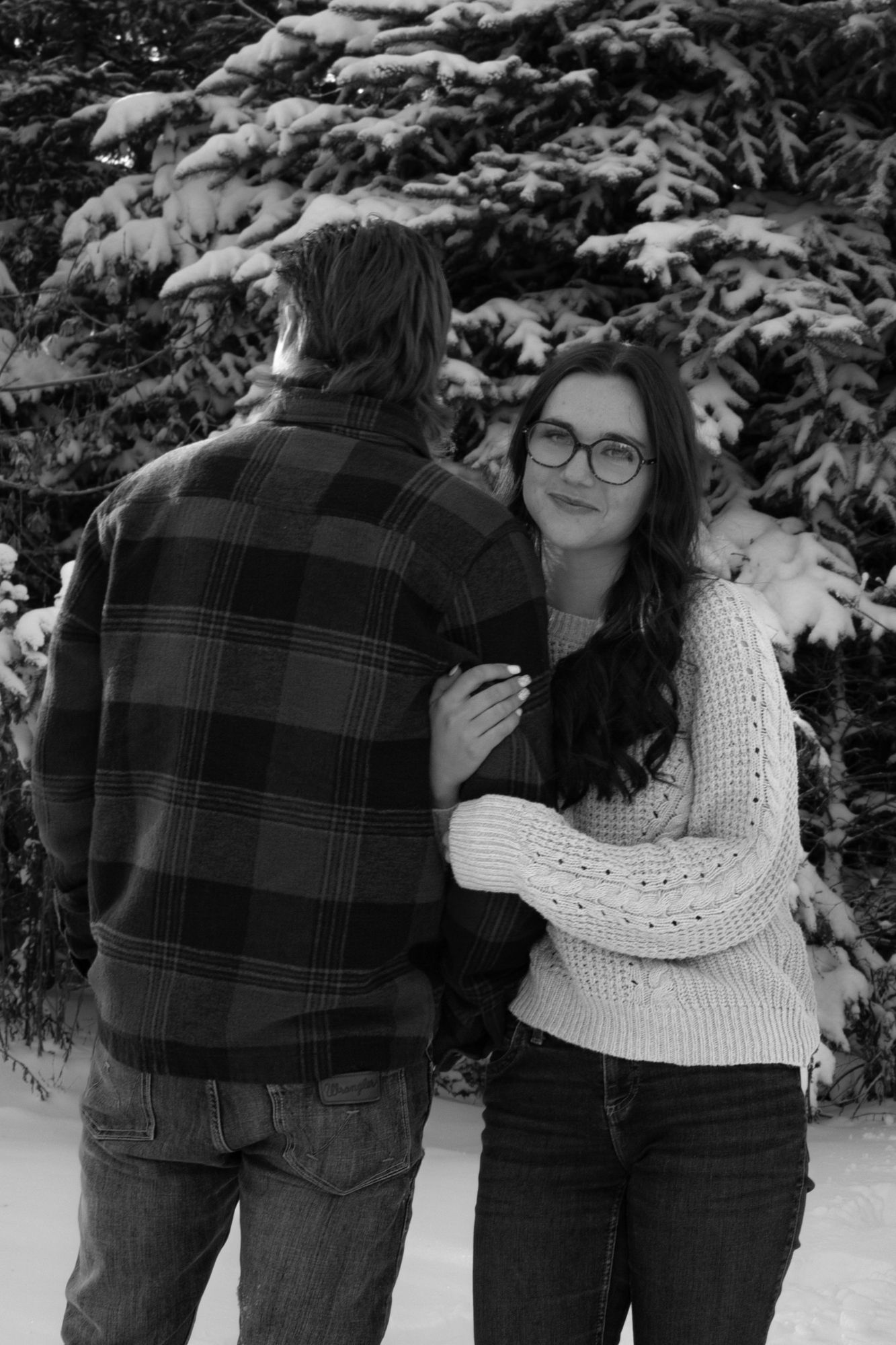 A couple embraces in the snow near snow-covered evergreen trees in a romantic winter scene.