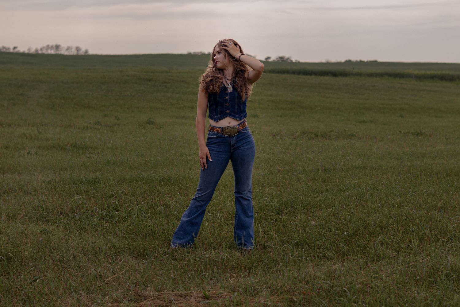 Woman in blue crop top and flare jeans poses in green field, hand on hat, cloudy sky backdrop.