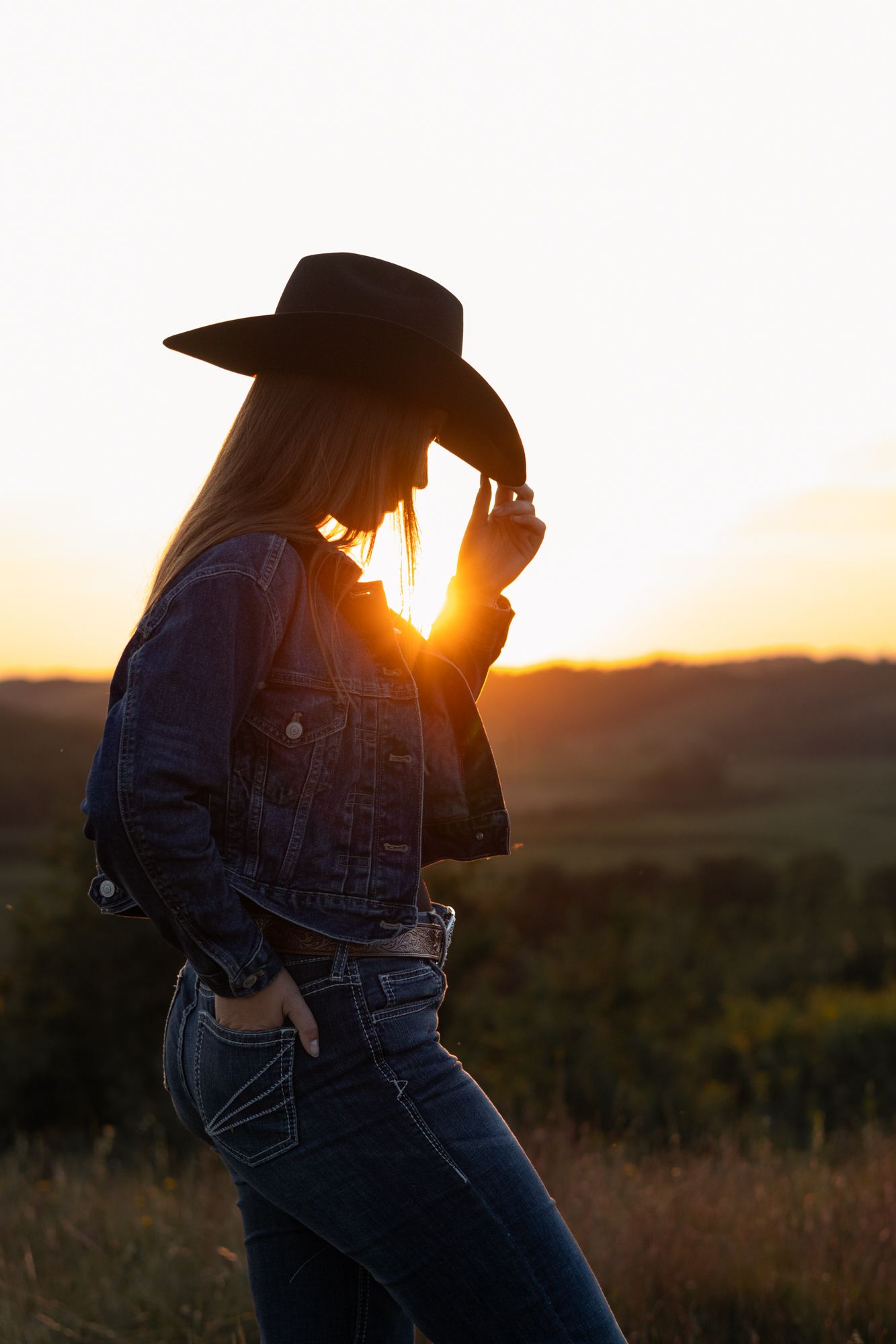 Silhouette of a woman in cowboy hat and denim jacket posing at golden sunset in a rural field.