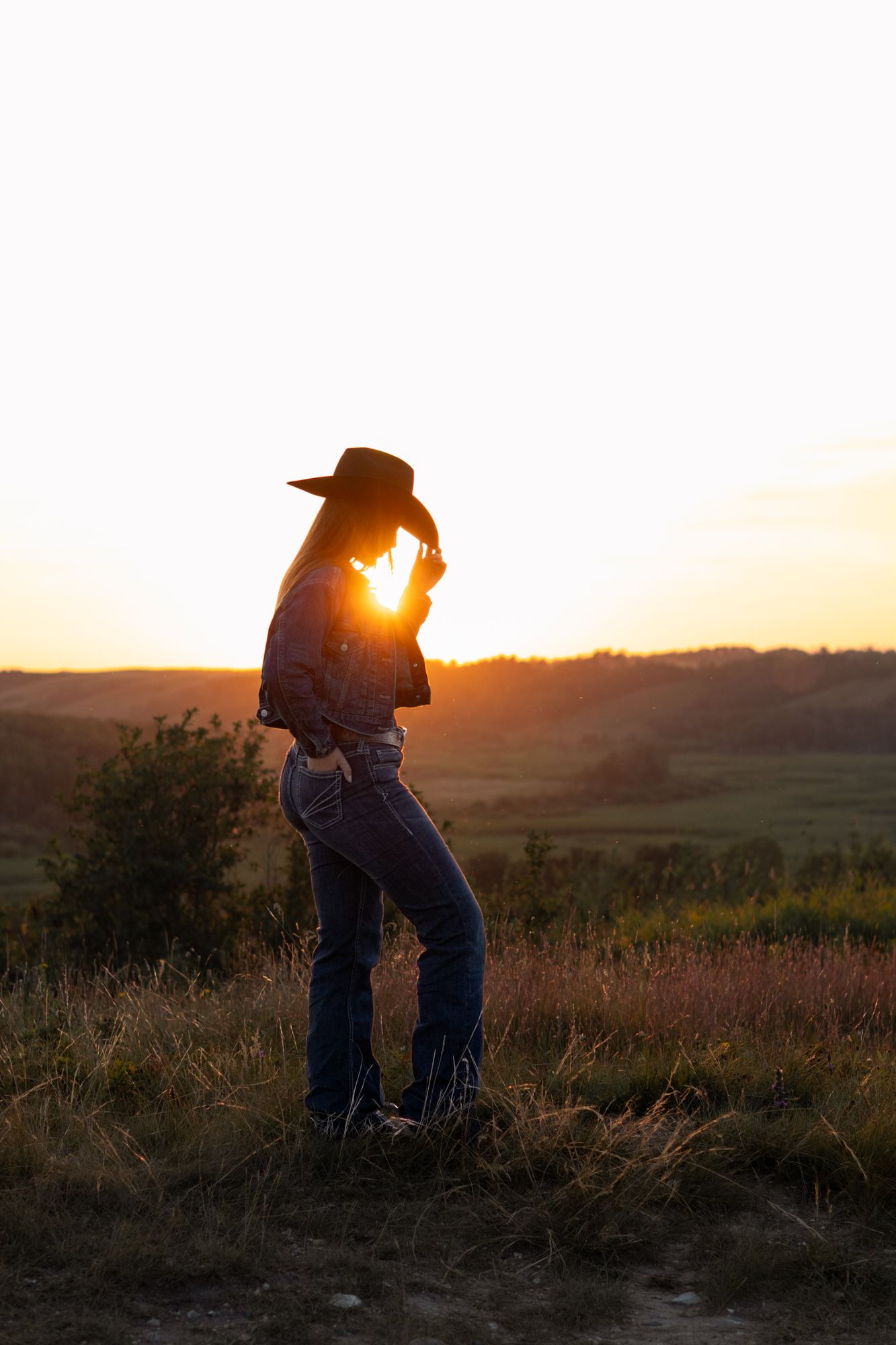 Woman in cowboy hat and jeans silhouetted against golden sunset over rolling countryside fields.